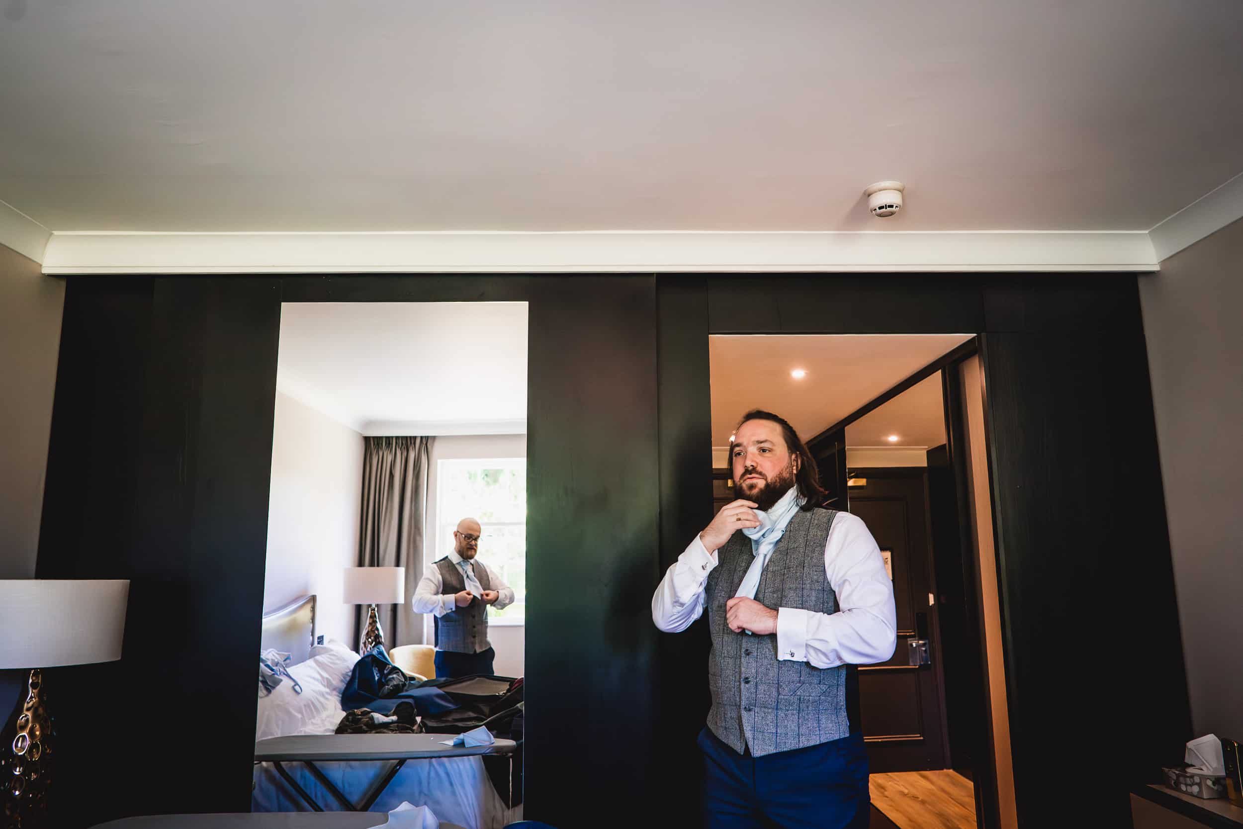 A man in a vest adjusts his tie in a hotel room with mirrored doors, reflecting him and a tidy bed.