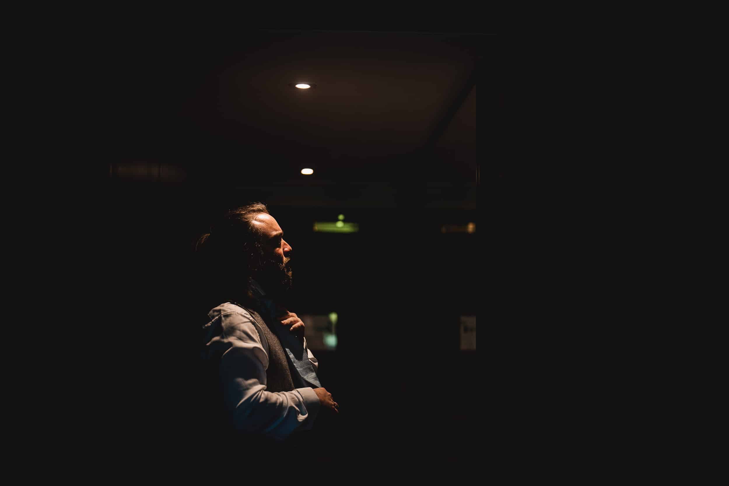 A man in a dimly lit room adjusts his tie while standing under a ceiling light, casting a shadow on the wall behind him.