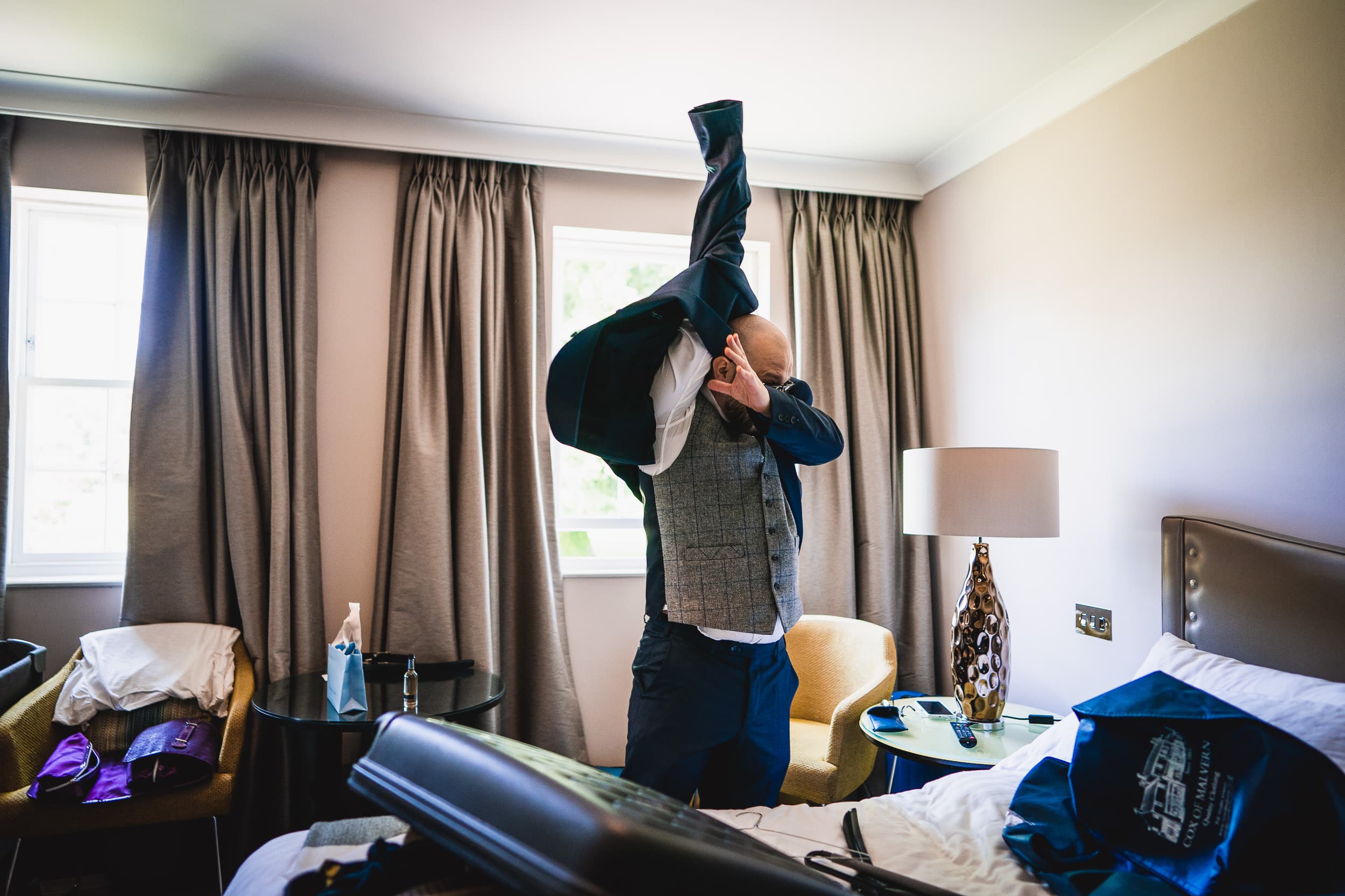 A man in a hotel room puts on a suit jacket. The room has curtains, a lamp, and a bed with various items on it.