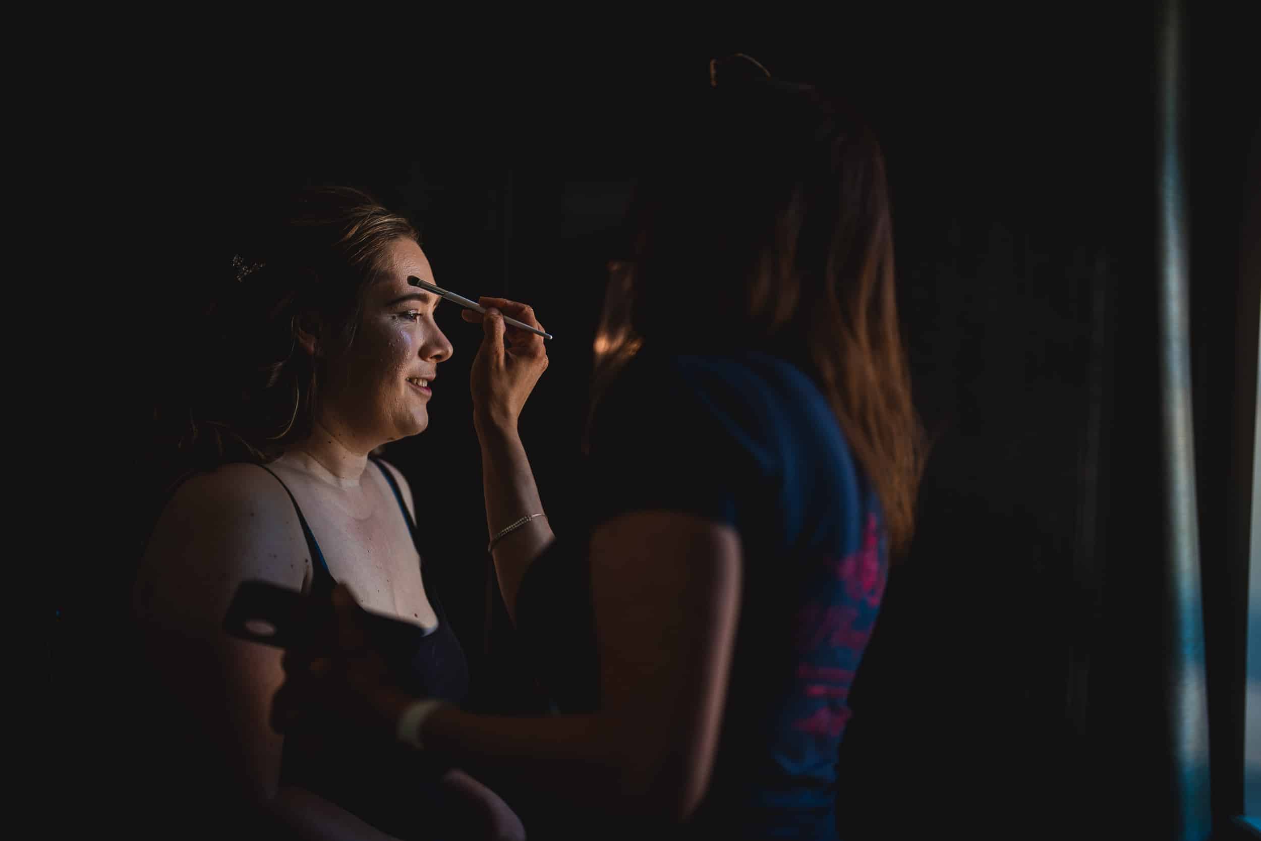 A woman applies makeup to another woman's eyebrows in a dimly lit setting.