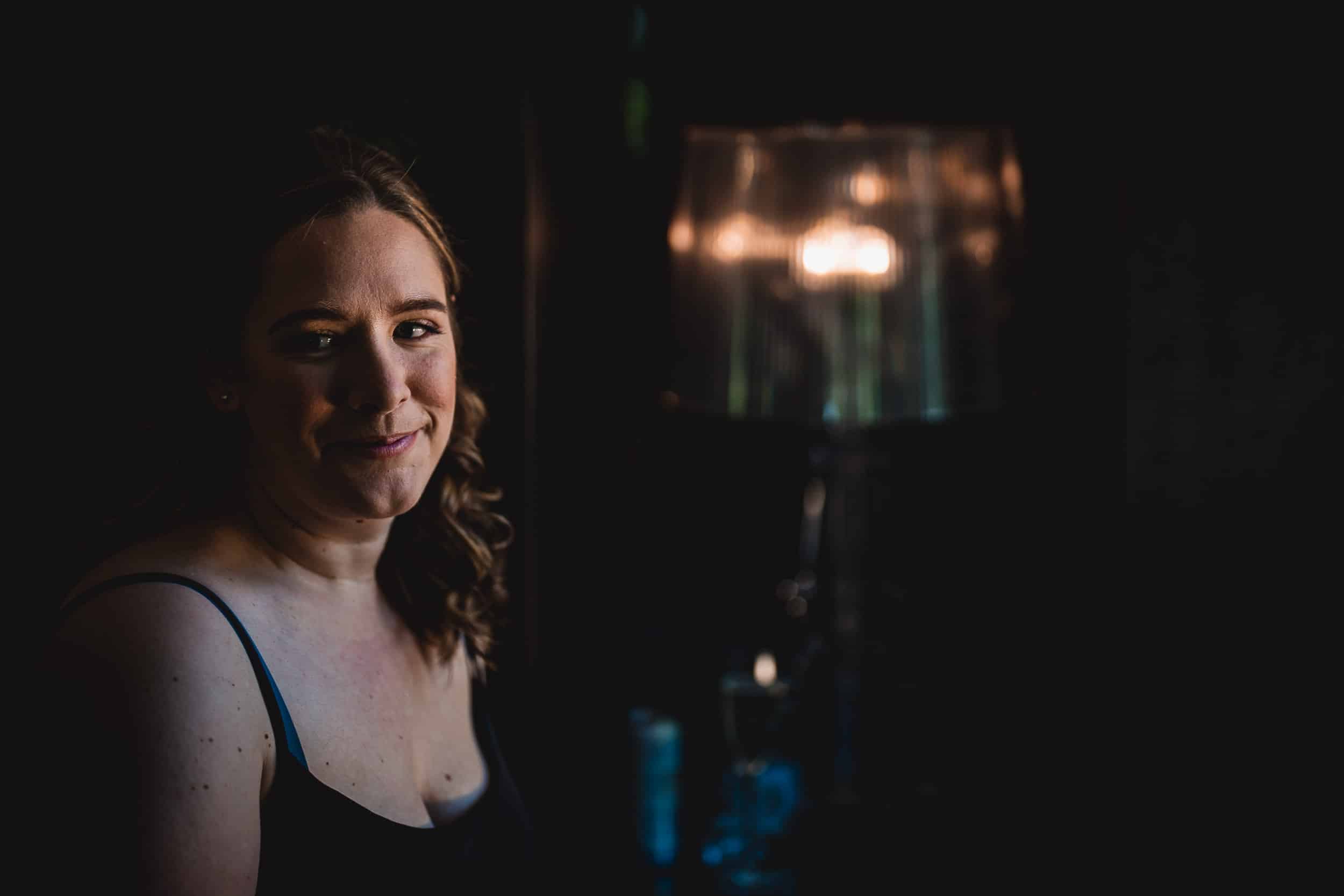 A woman with long hair sits in a dimly lit room next to a glowing lamp, wearing a sleeveless top.