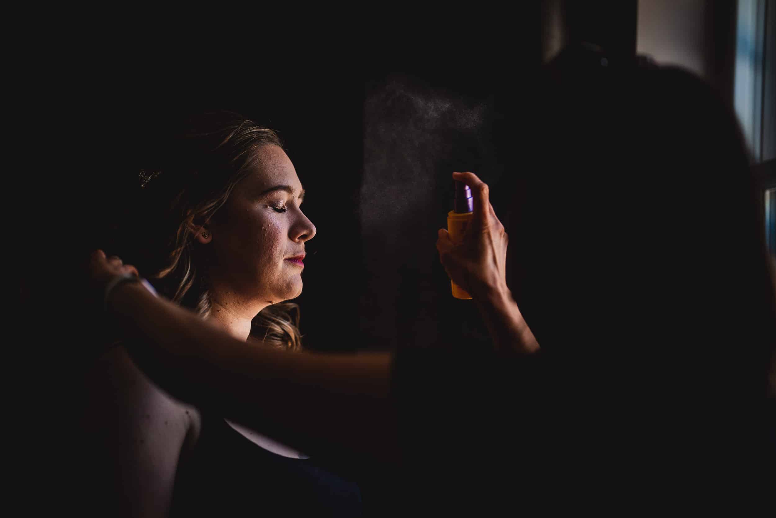 A woman with closed eyes is having her makeup set with a spray by another person in a dimly lit room.