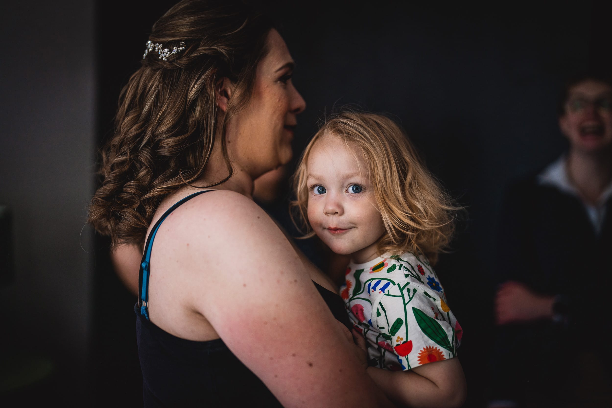 A woman holding a child with blonde hair. The child is wearing a floral patterned shirt and looking at the camera with a slight smile.