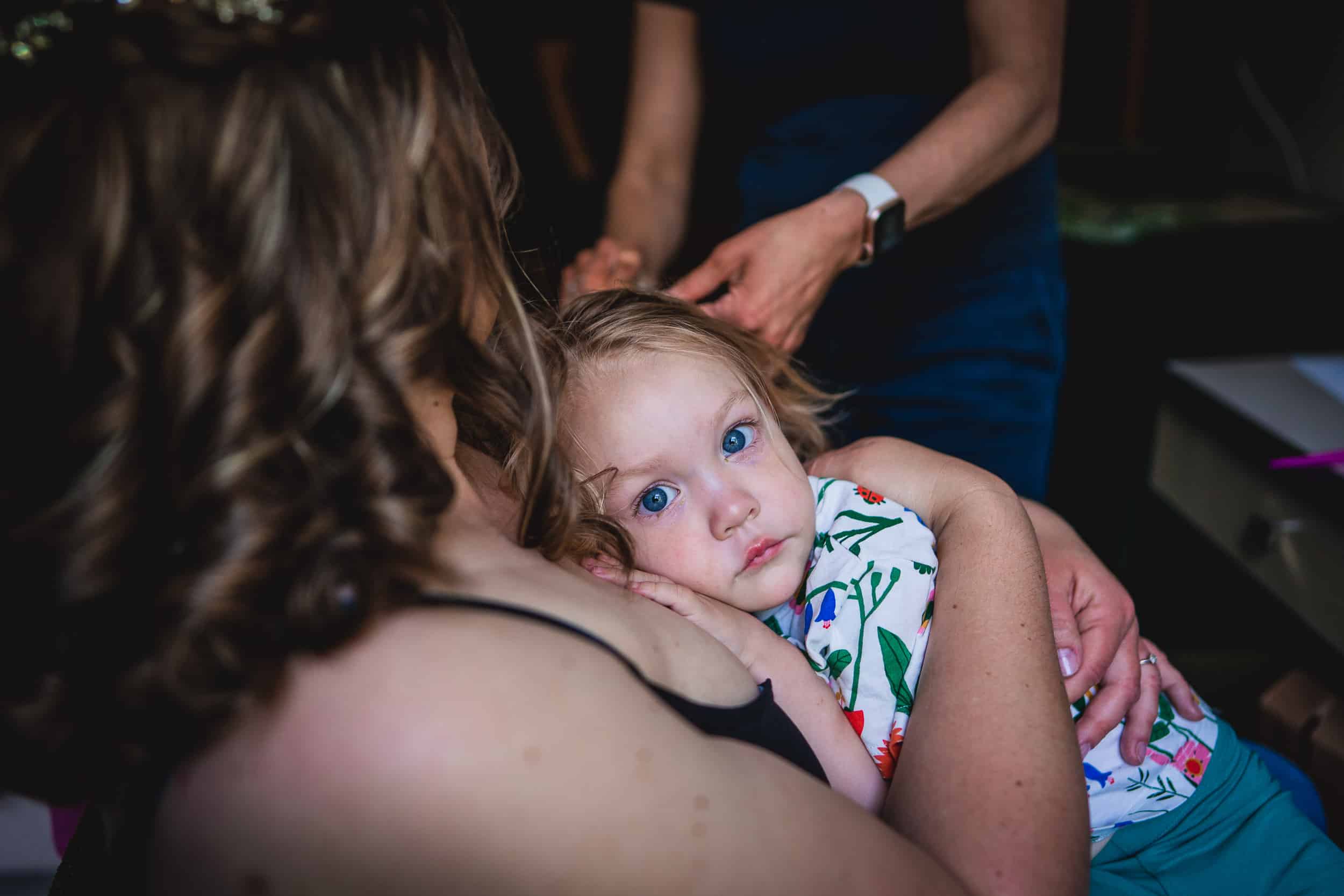 A child with blue eyes is resting in an adult's arms. The child wears a floral-patterned outfit, and an adult's hands seem to be adjusting the child's hair.