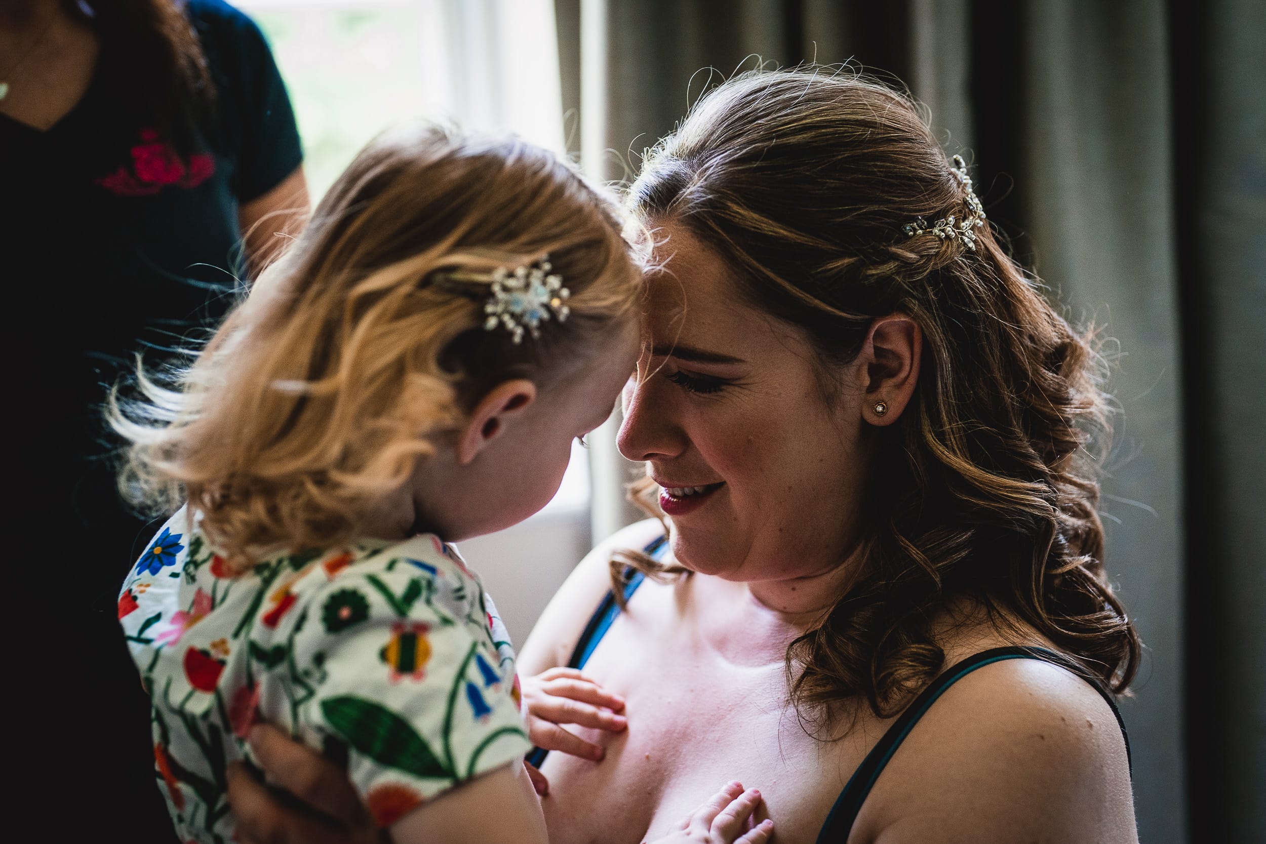 A woman and a young child with flower hairpins share a close, affectionate moment indoors.