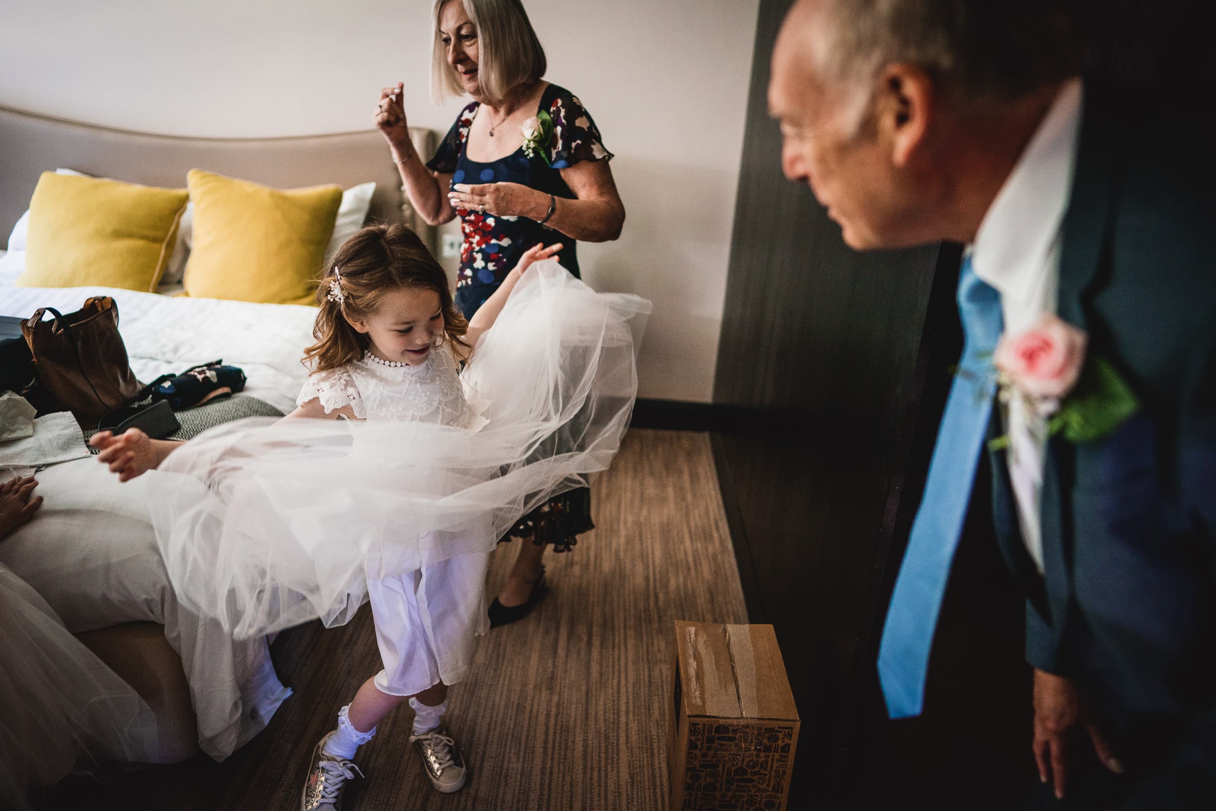 A young girl plays with her dress in a room, surrounded by smiling adults. A man in a suit looks on, and a woman in a floral dress stands nearby.