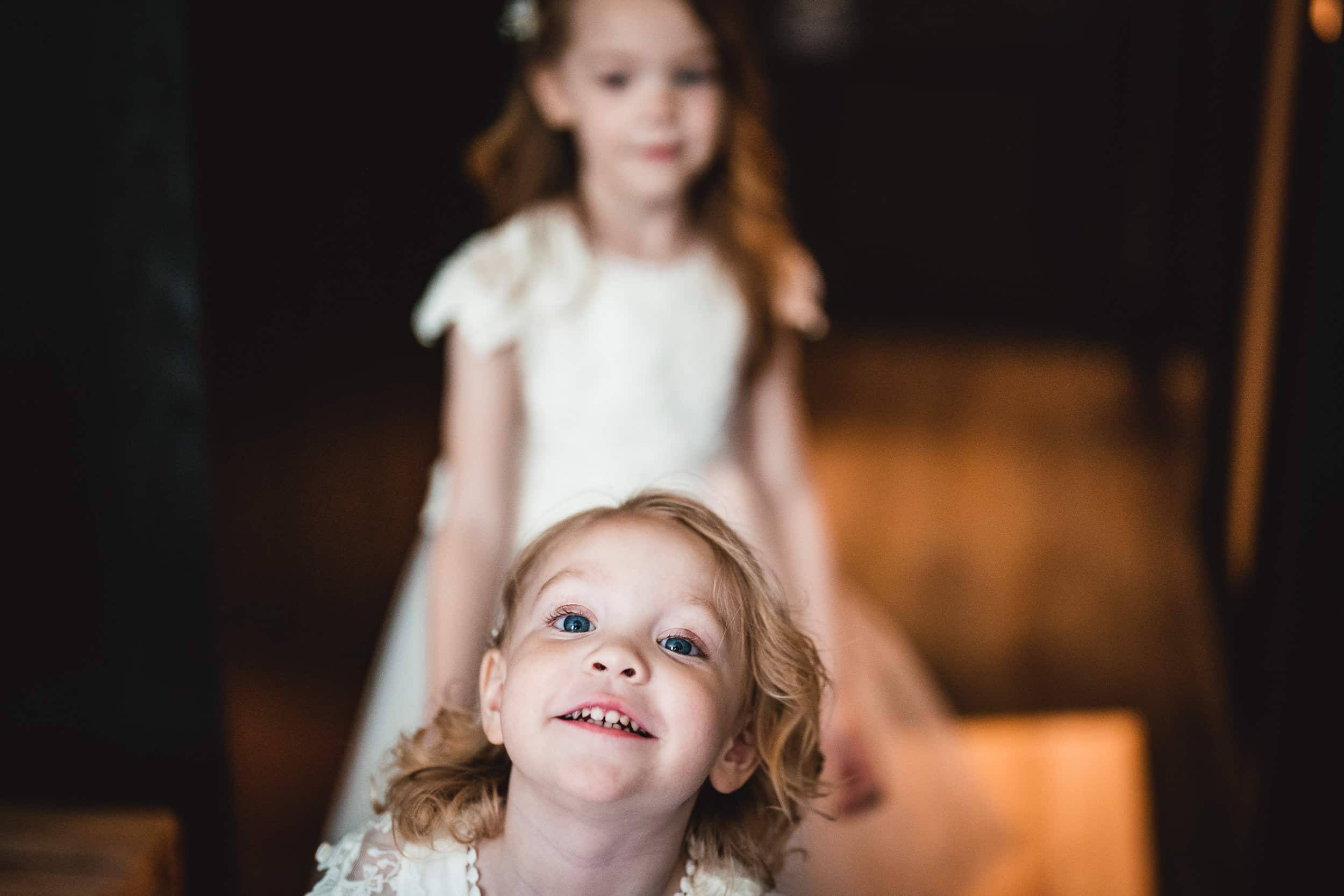 Two young girls wearing white dresses are indoors. The girl in the foreground is looking up and smiling, while the other stands behind her.