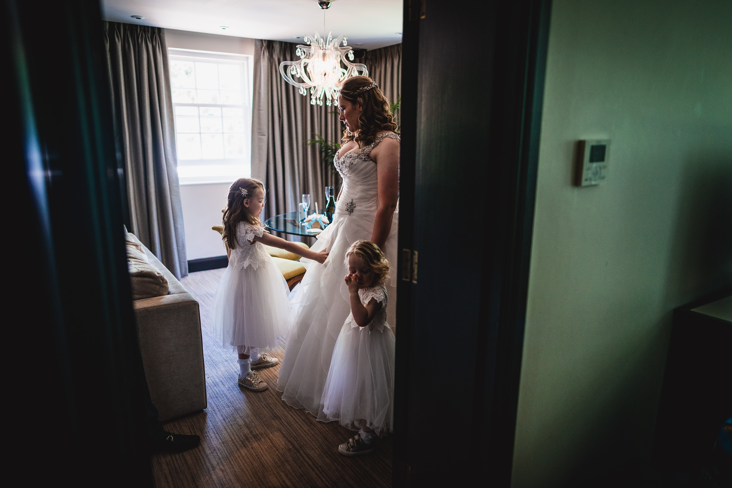 A bride and two young girls in white dresses stand in a room with a chandelier and a window with curtains.