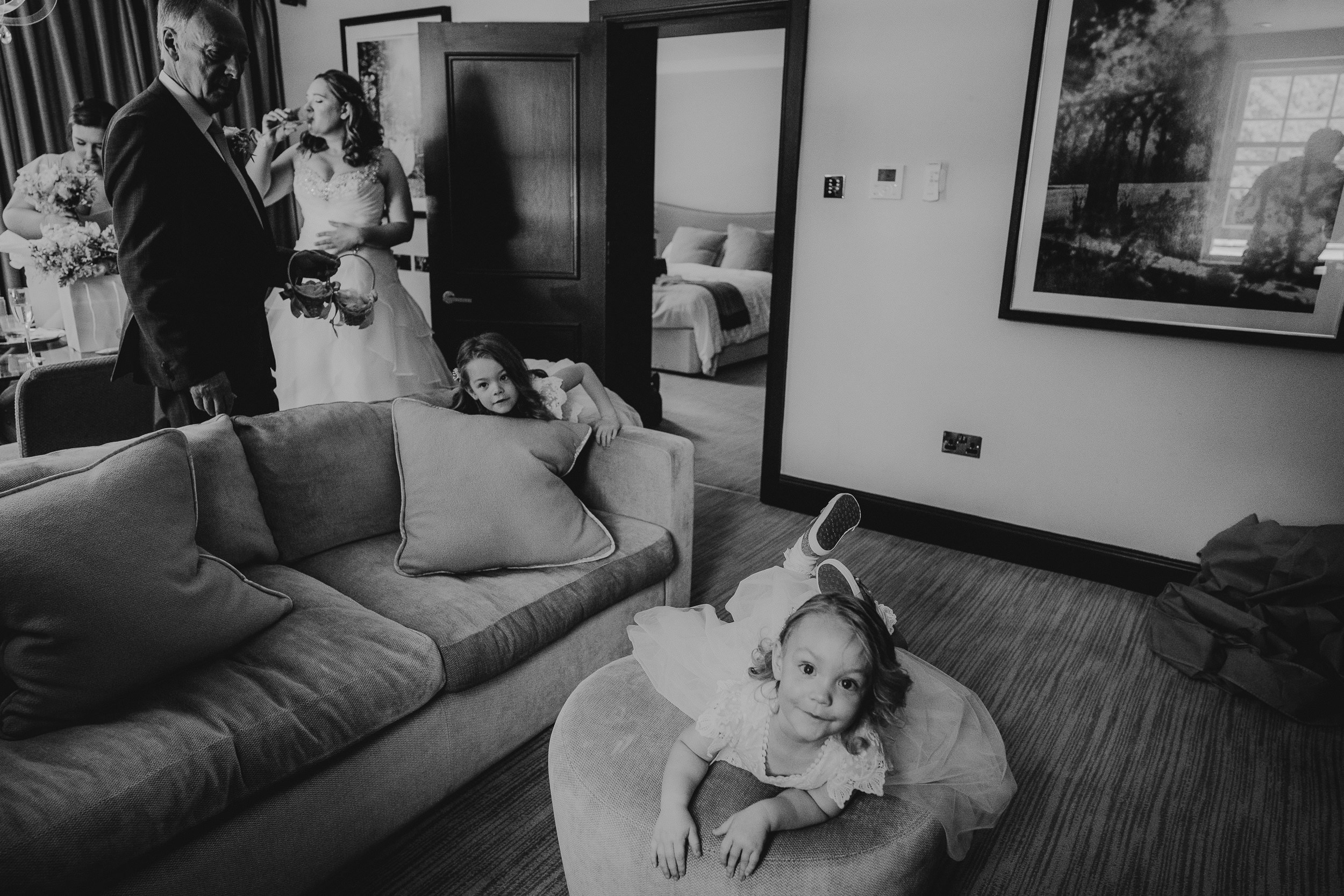 A black-and-white image of a family in a living room. A child lies on an ottoman, another sits on a couch, and two adults stand near a doorway, with one holding a bouquet.