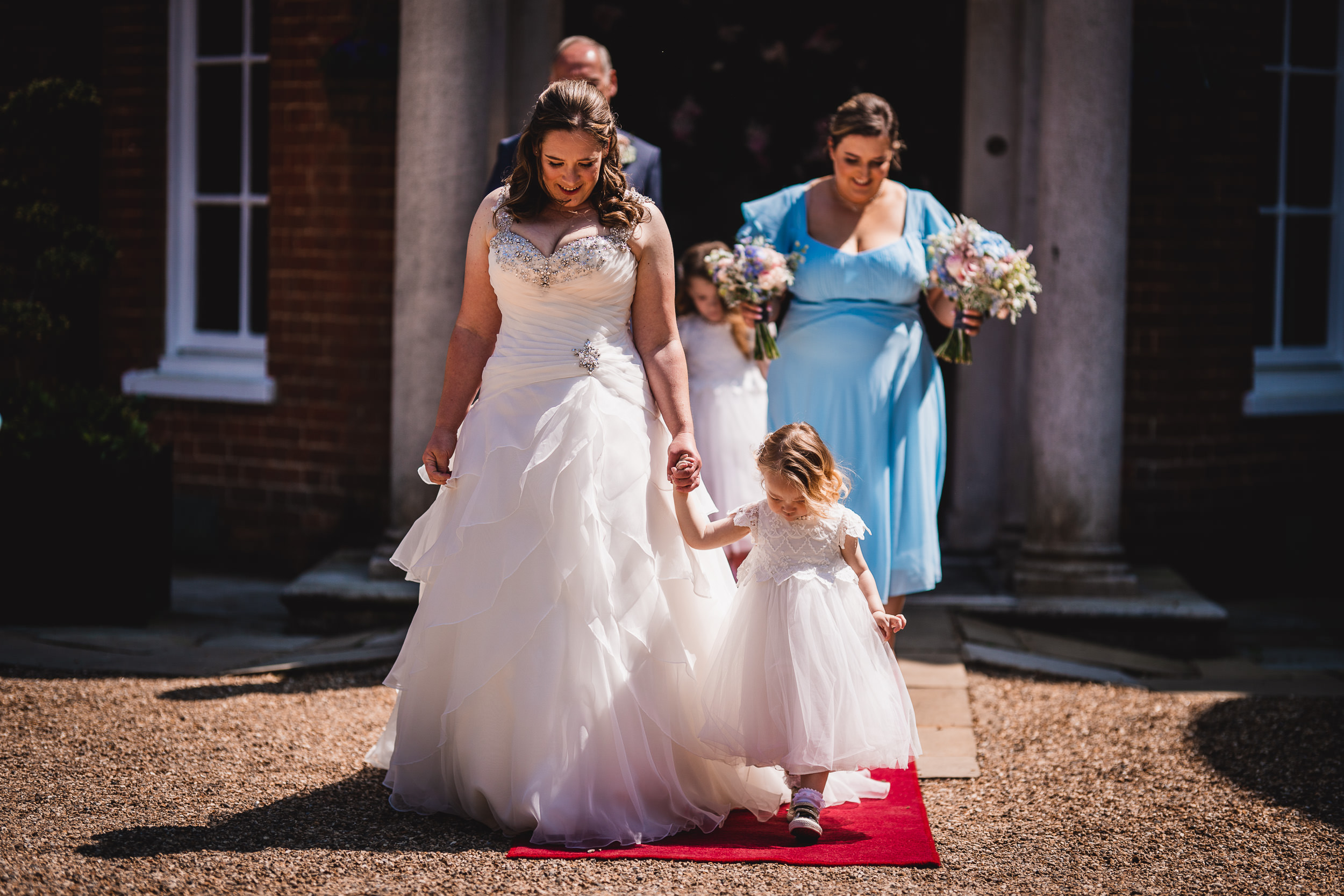 A bride in a white gown walks down an outdoor path, holding hands with a small child. Two women and another child follow, with one in a blue dress holding flowers.