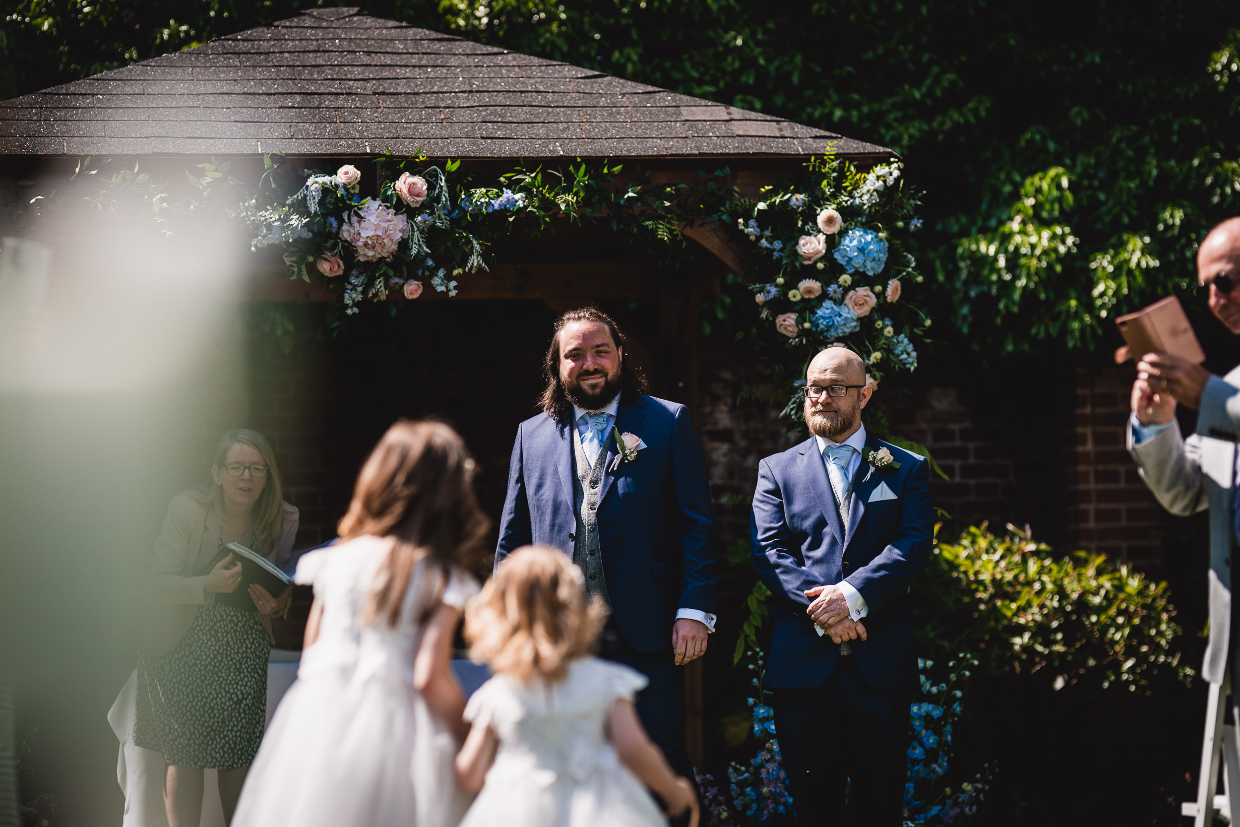 A groom and a groomsman stand under a gazebo adorned with flowers while two flower girls walk towards them at an outdoor wedding.