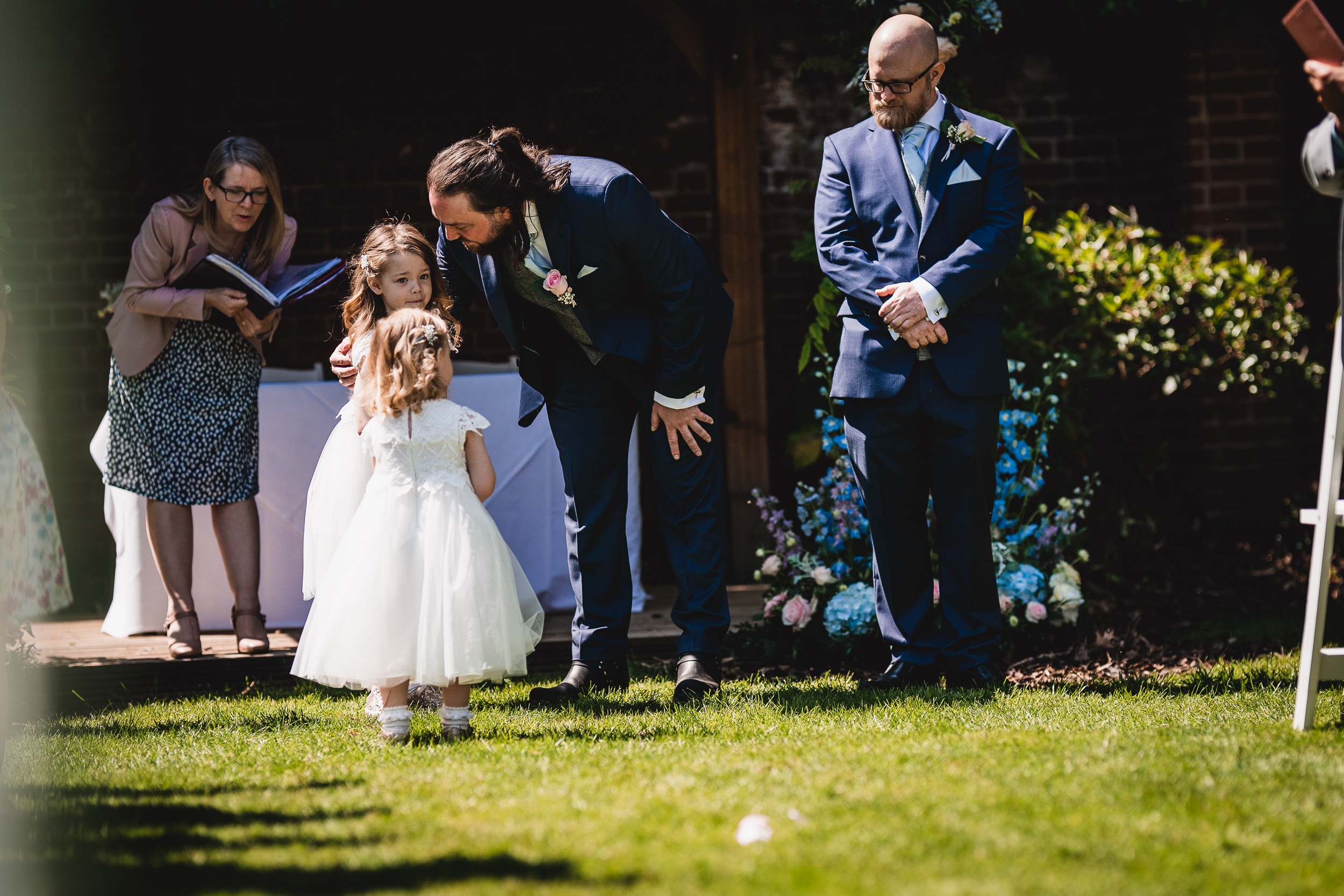 A man in a suit bends down to speak with two young girls in white dresses at an outdoor ceremony. A suited man stands nearby, and a woman is holding a book in the background.