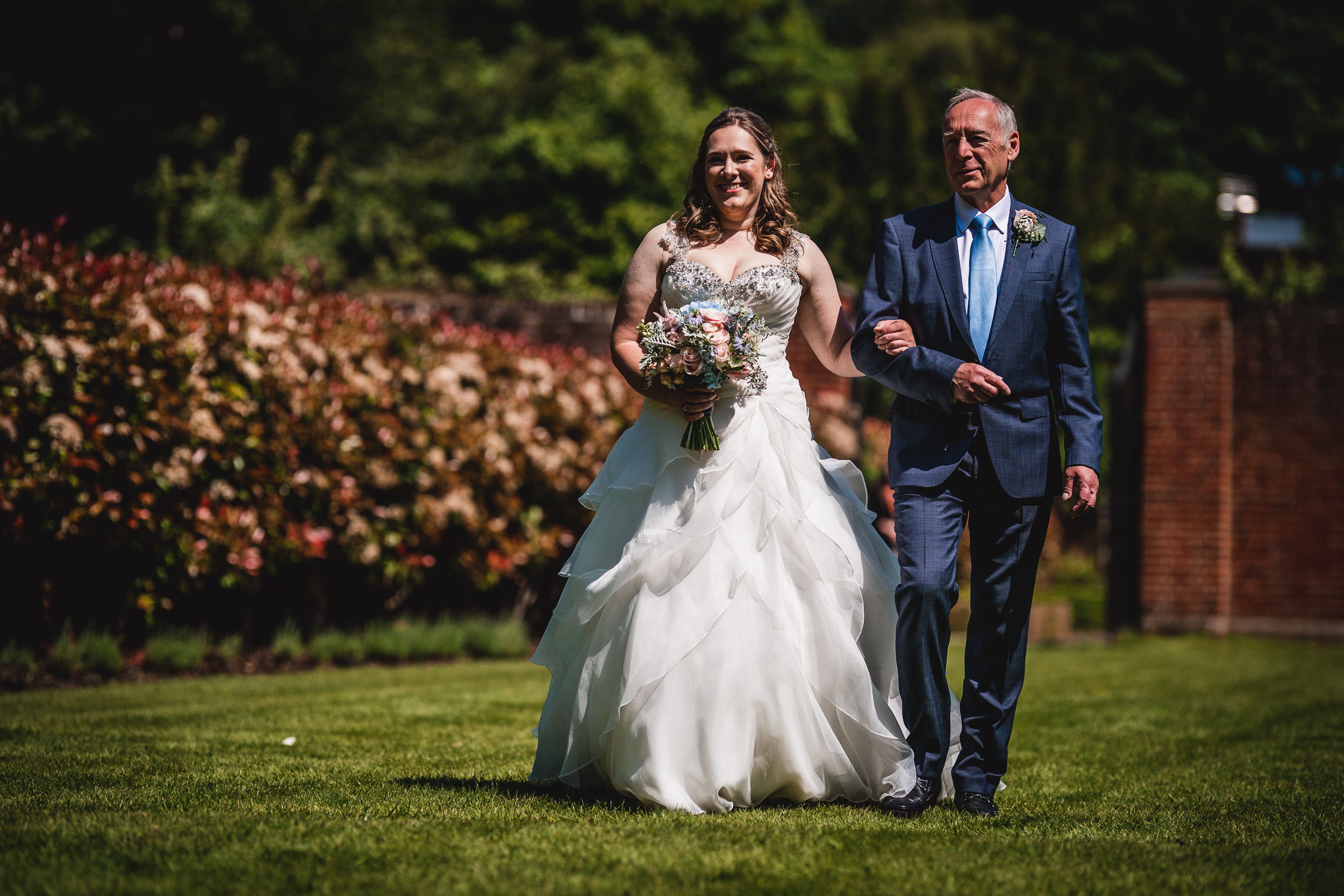 A bride in a white gown holds a bouquet, walking beside a man in a suit on a sunny day with greenery and a brick wall in the background.