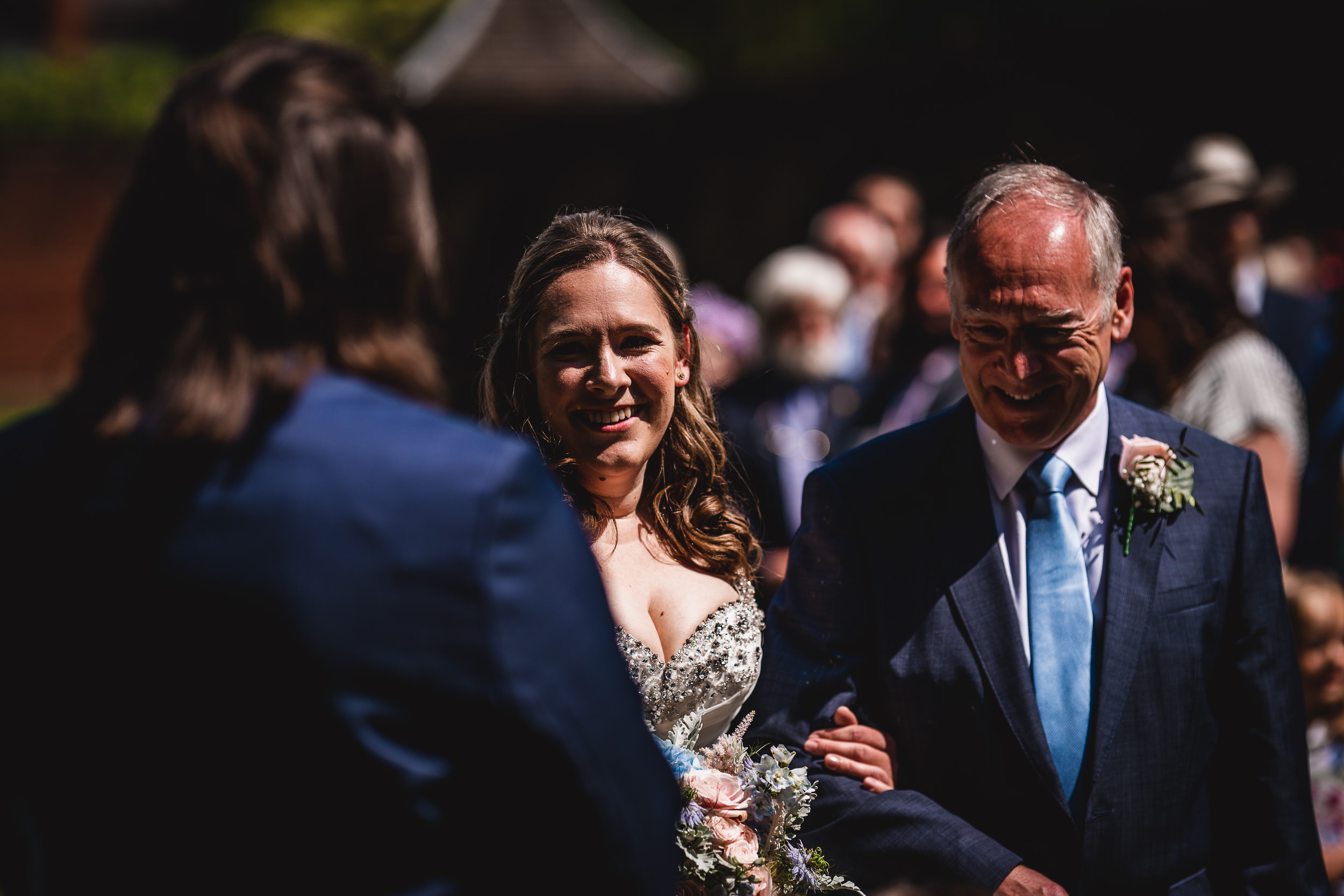 A bride walks down the aisle with an older man in a suit, surrounded by onlookers, during an outdoor ceremony.