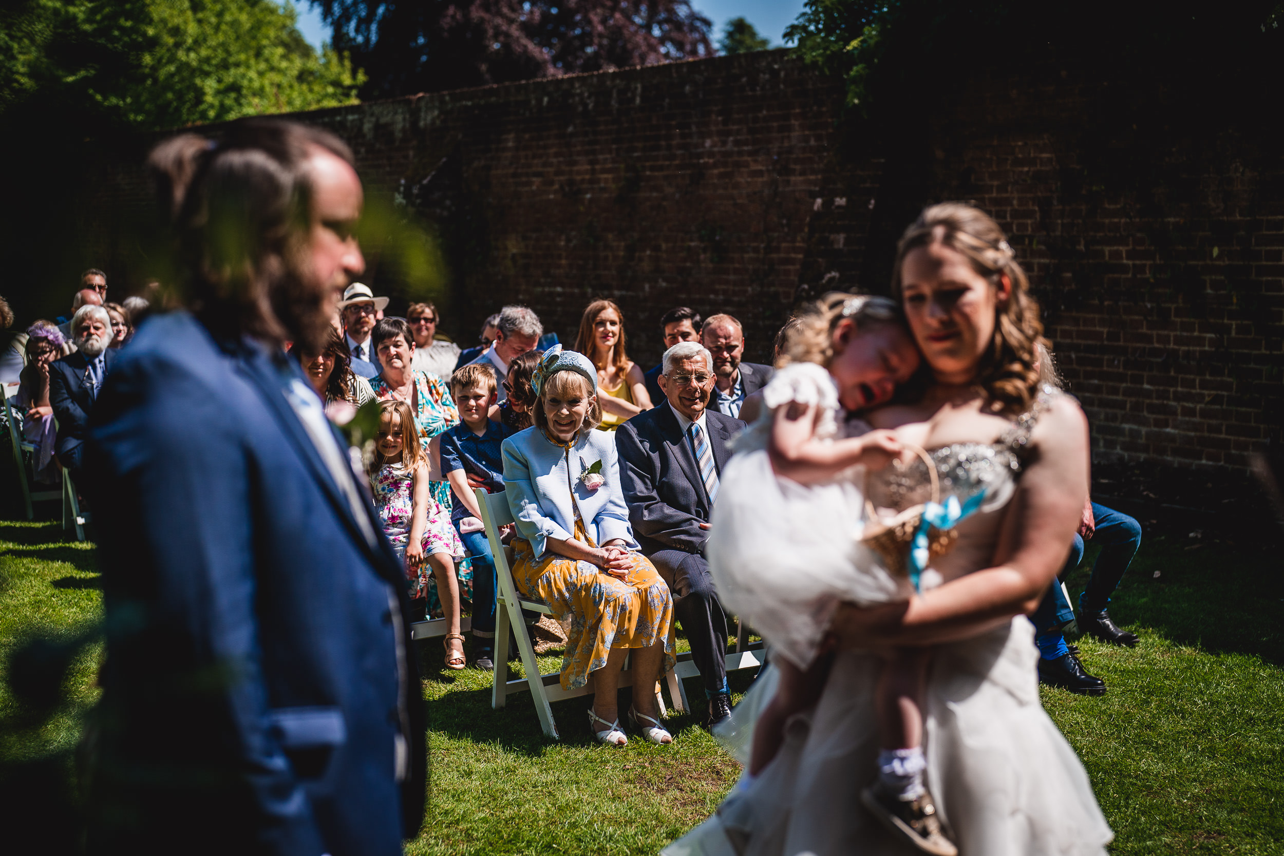 A woman holds a child at an outdoor wedding ceremony. Guests are seated in rows, watching the event. A man stands towards the front. Sunlight filters through the trees.