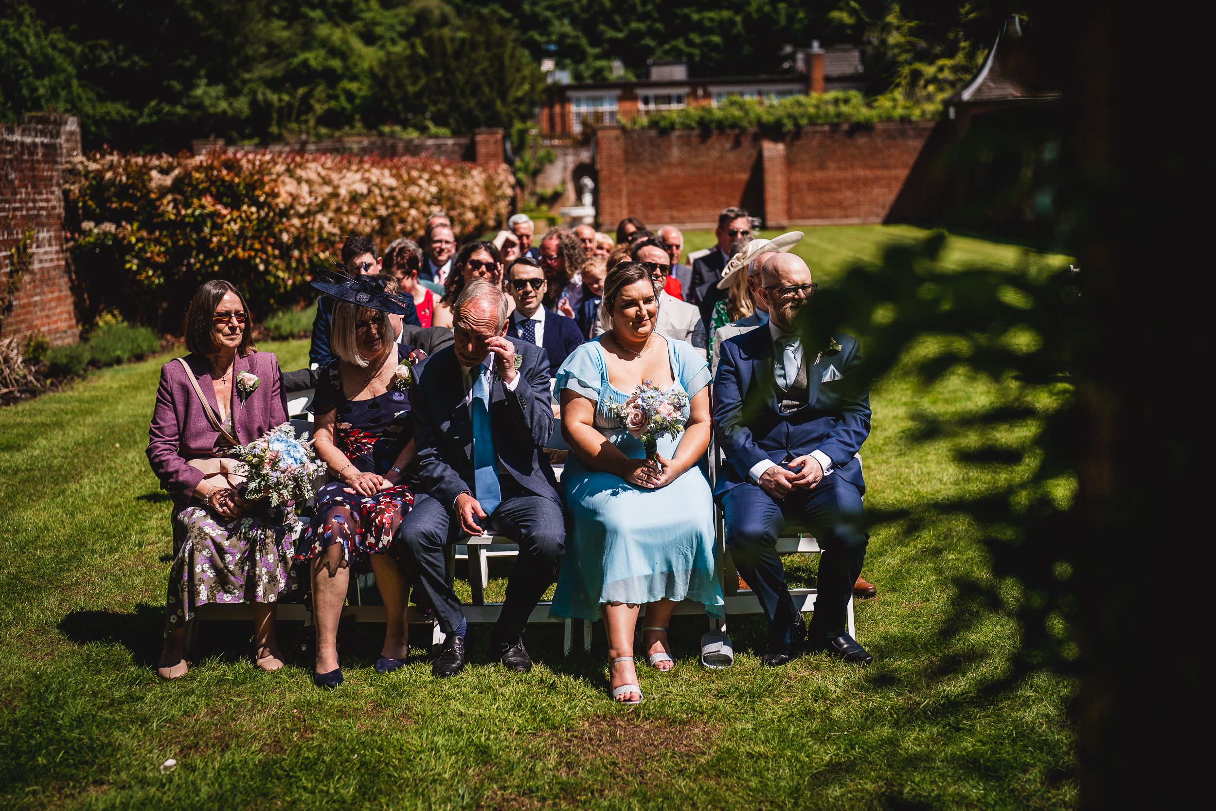 Guests seated outdoors at a wedding, with some holding floral bouquets. They are dressed in formal attire on a sunny day, surrounded by greenery and brick walls.