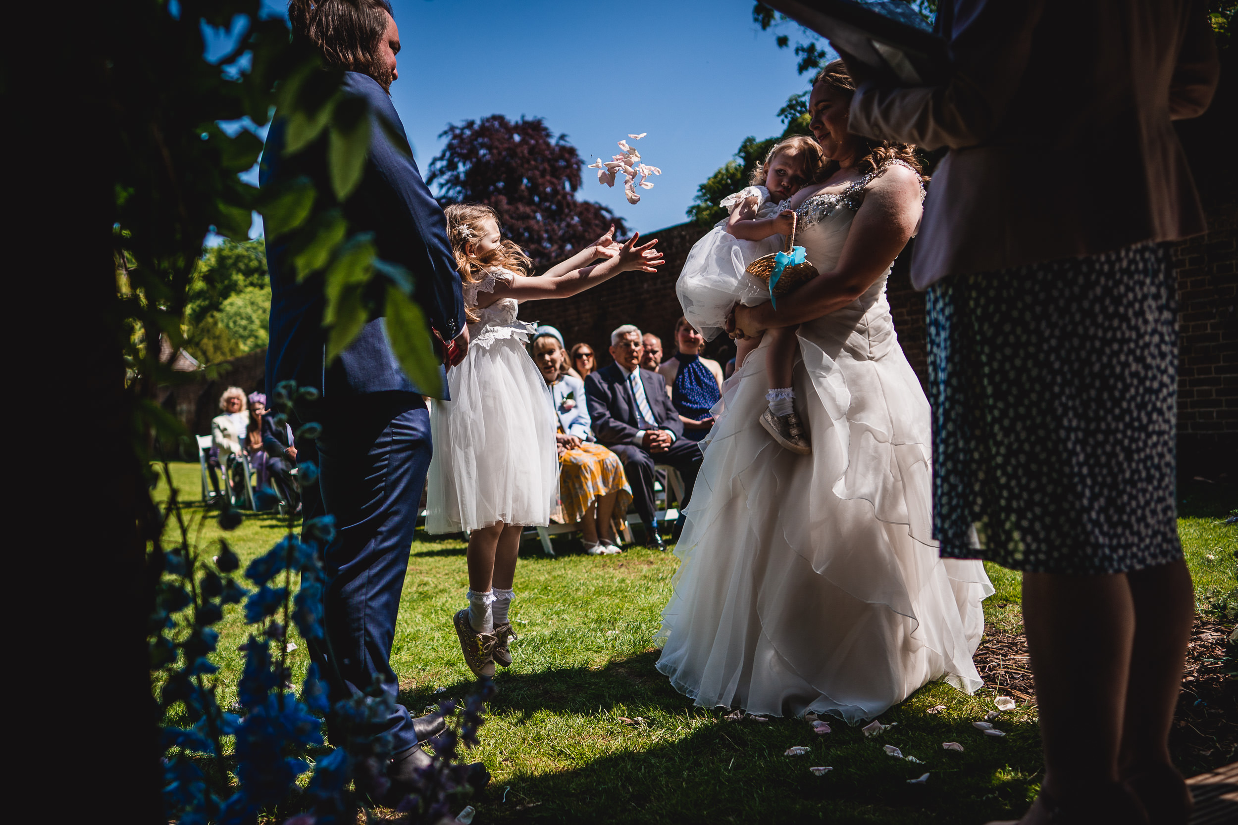 A bride holding a child interacts with a young girl throwing flower petals in an outdoor wedding setting with seated guests.