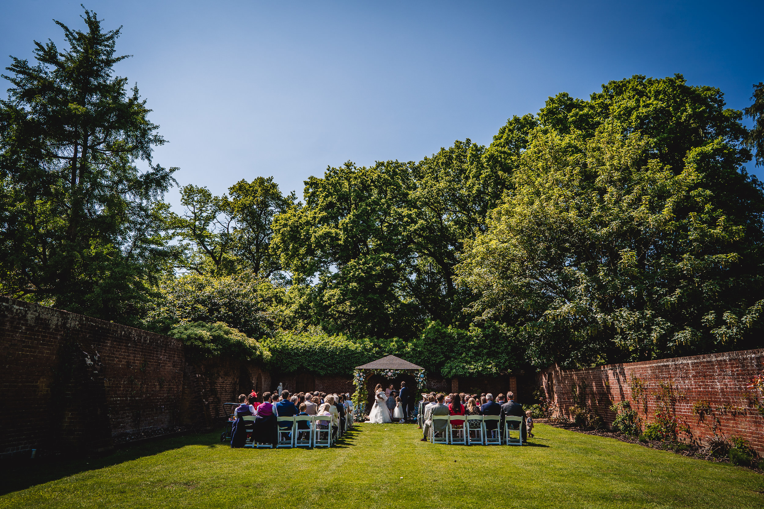 Outdoor wedding ceremony with guests seated on either side of an aisle in a garden setting, surrounded by brick walls and trees under a clear blue sky.