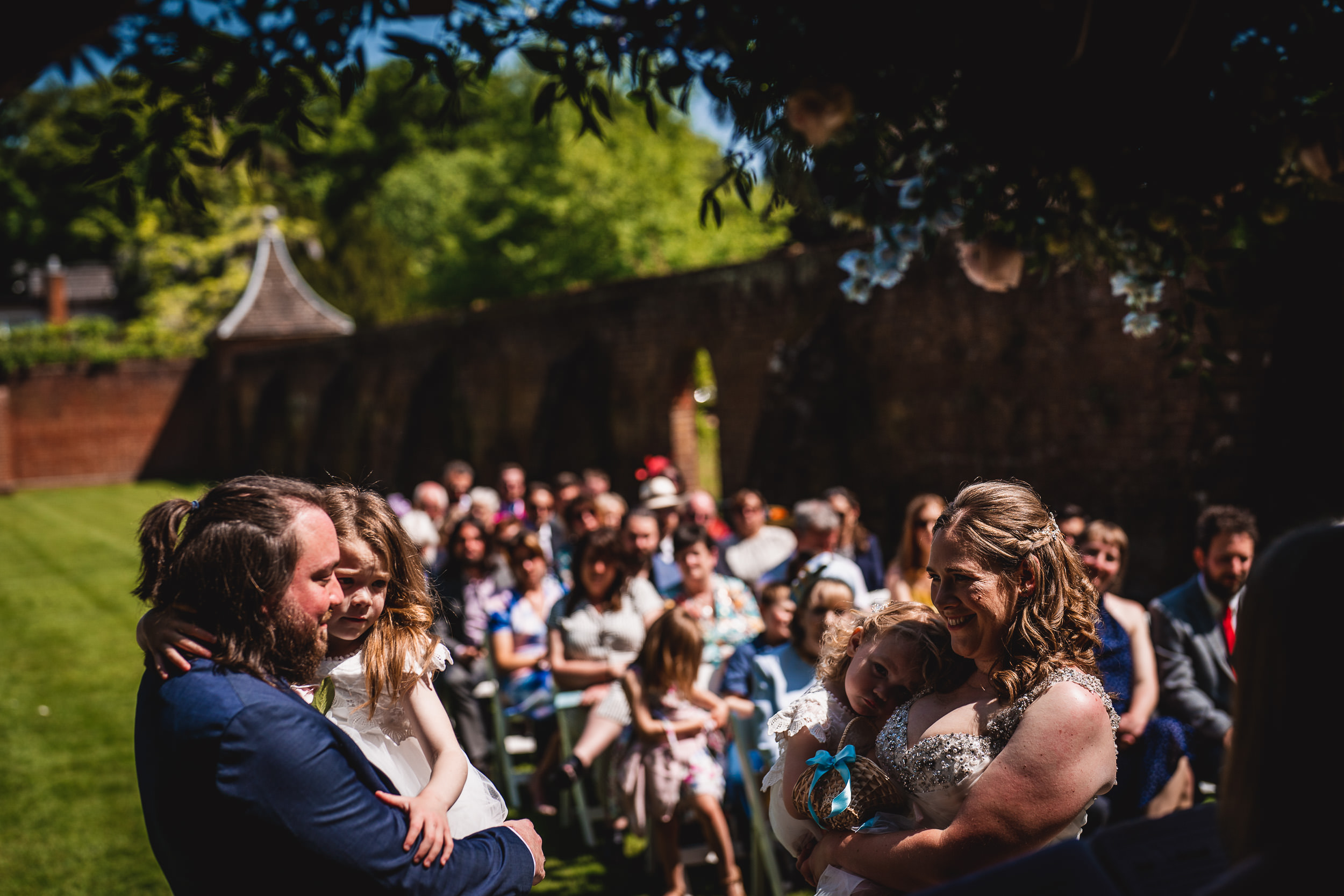 A couple stands at an outdoor wedding ceremony, each holding a child. Guests are seated in rows behind them, with trees and a brick wall in the background.