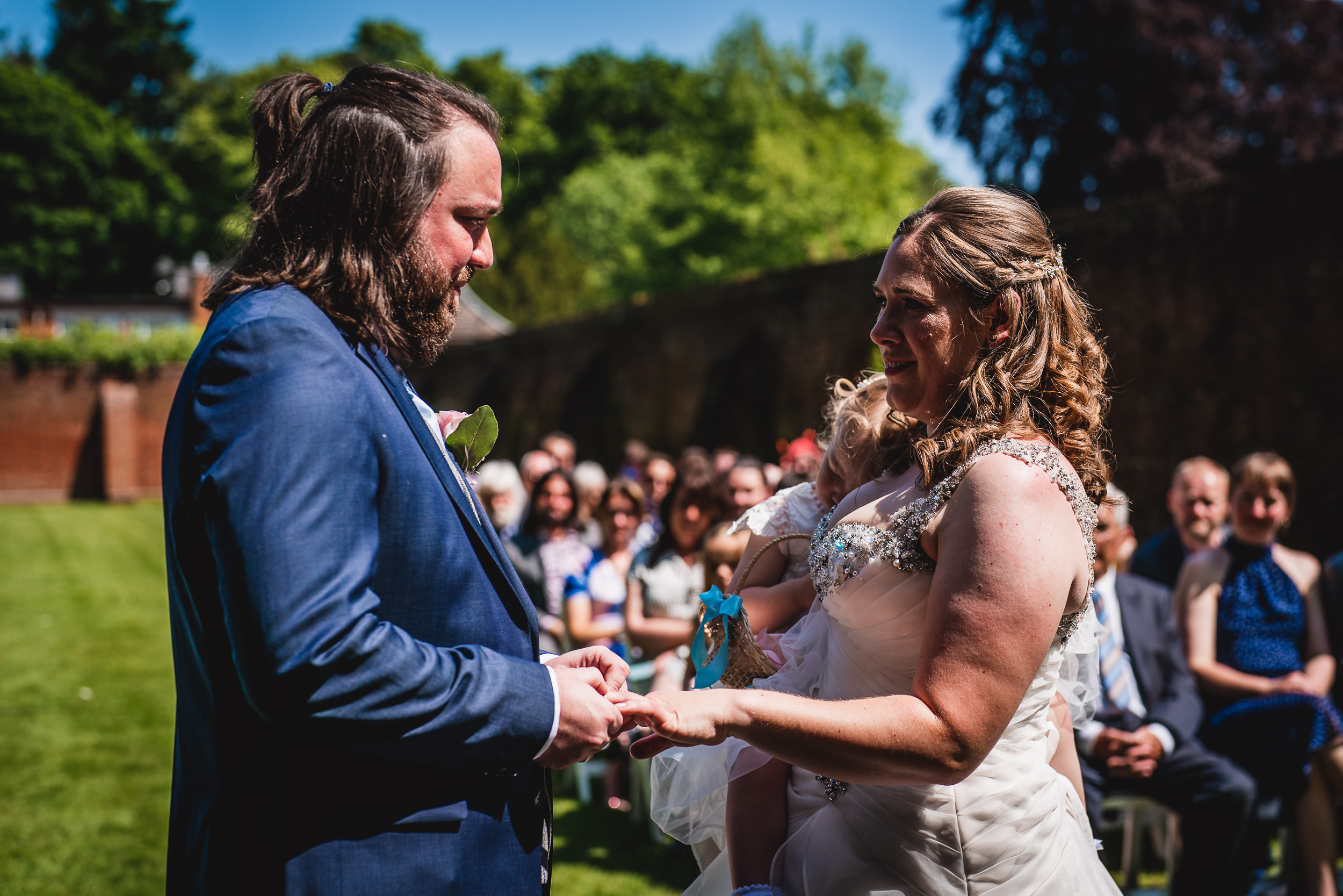 A couple exchanges rings during an outdoor wedding ceremony, with seated guests watching.