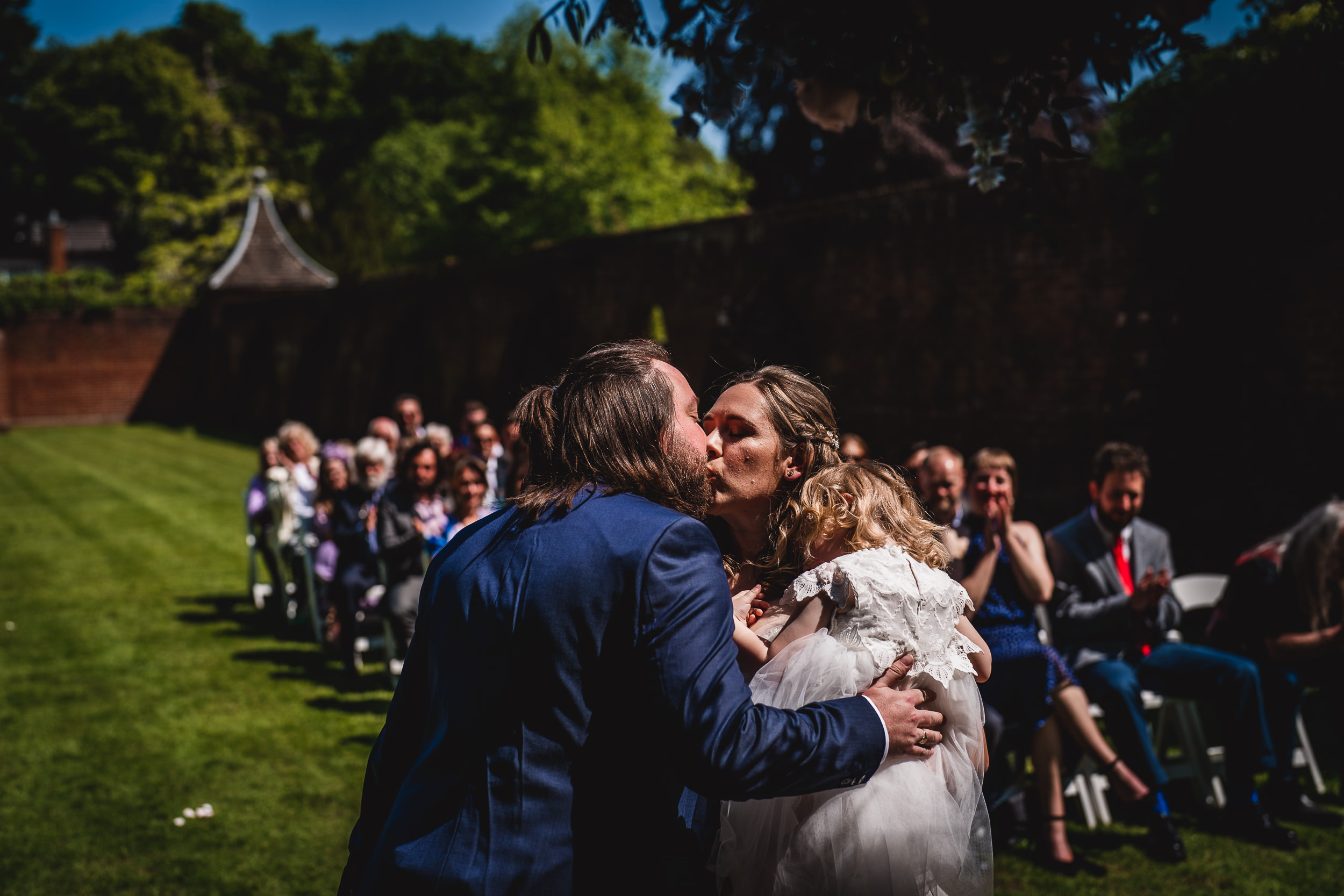 A couple kisses while holding a child during an outdoor wedding ceremony, with seated guests watching in the background.