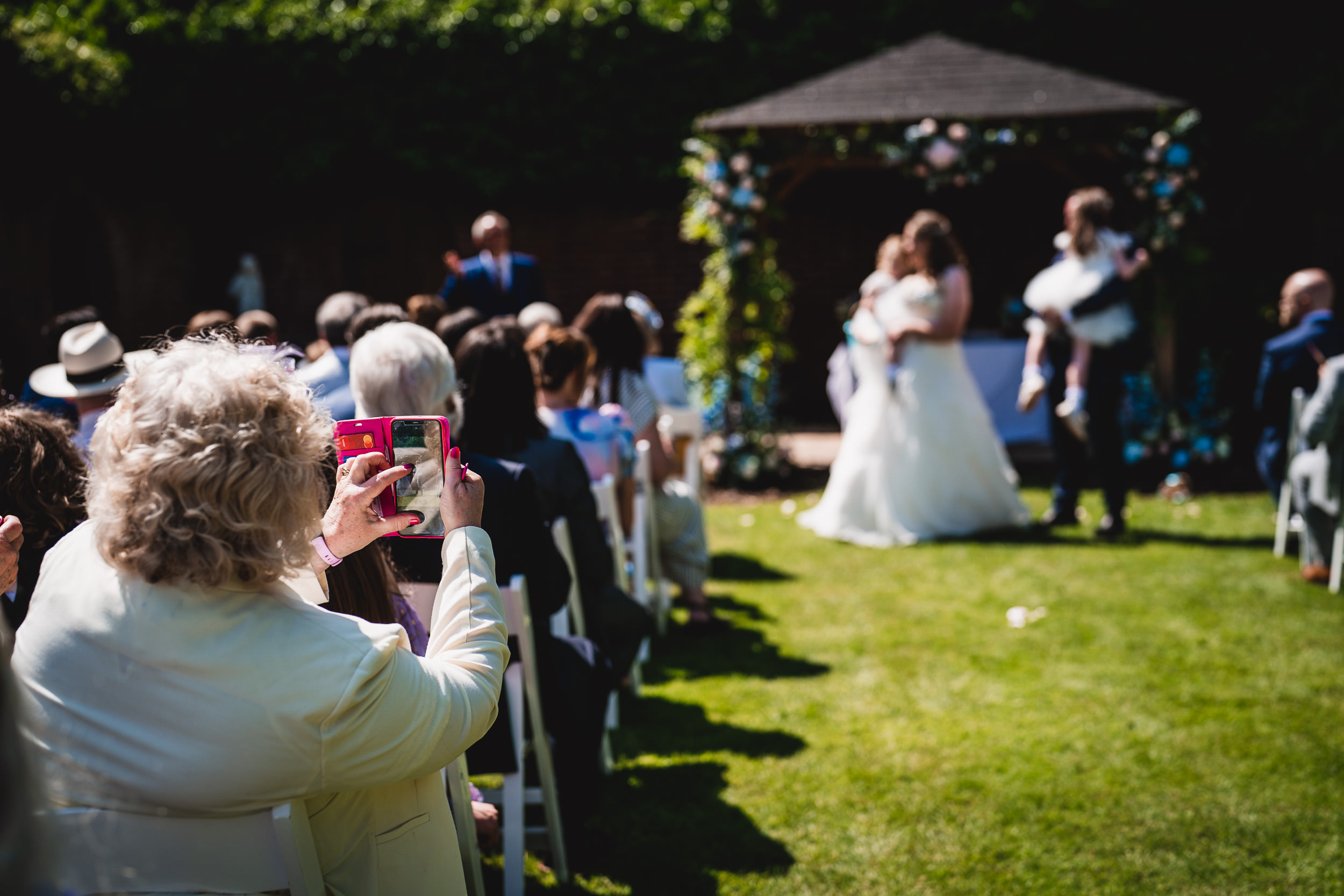 A person takes a photo with a phone at an outdoor wedding ceremony. The bride and groom stand under a gazebo in the background. Guests are seated on either side of the aisle.