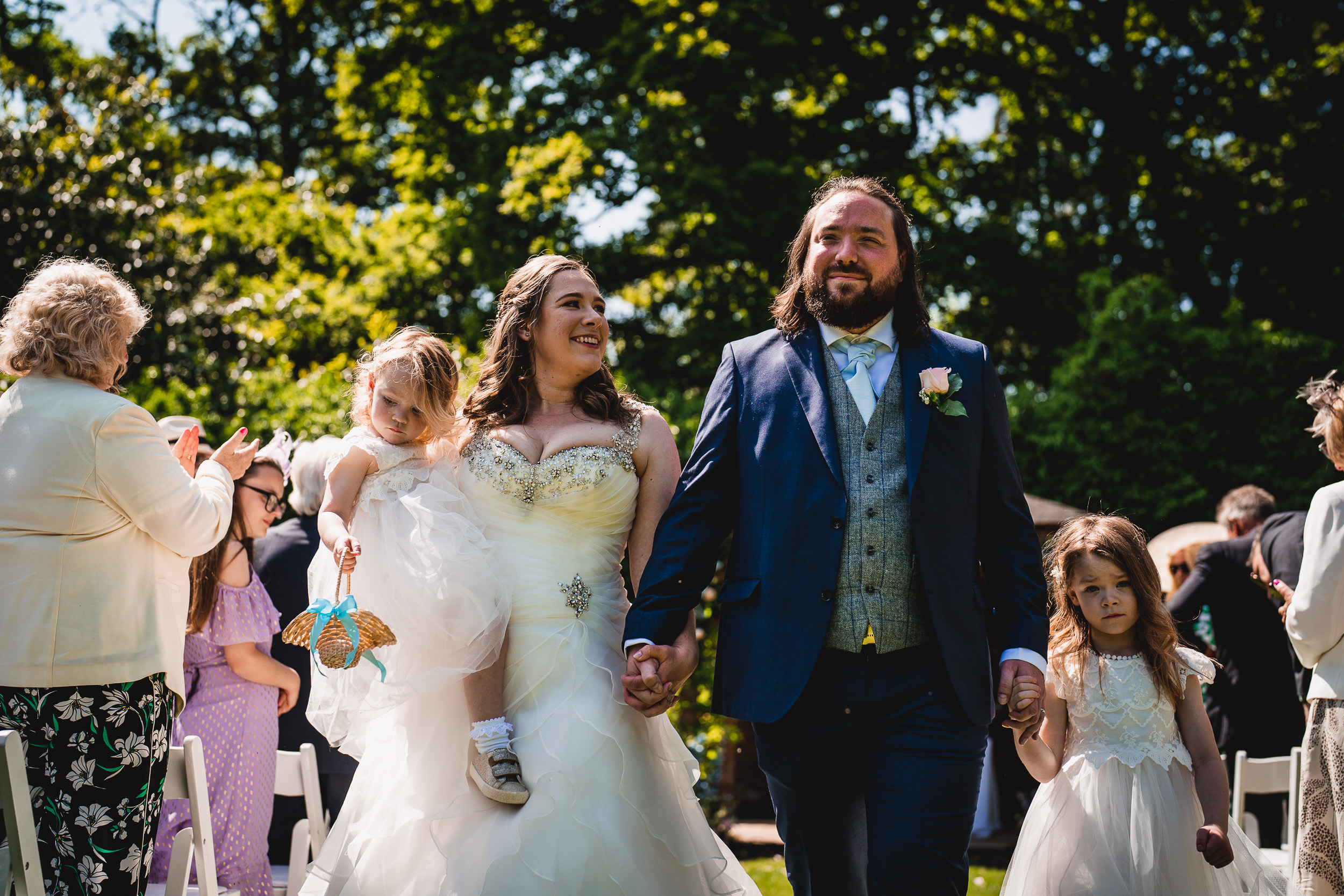 A newlywed couple walks down an aisle in an outdoor setting, holding hands with two young children. Guests clap in the background.