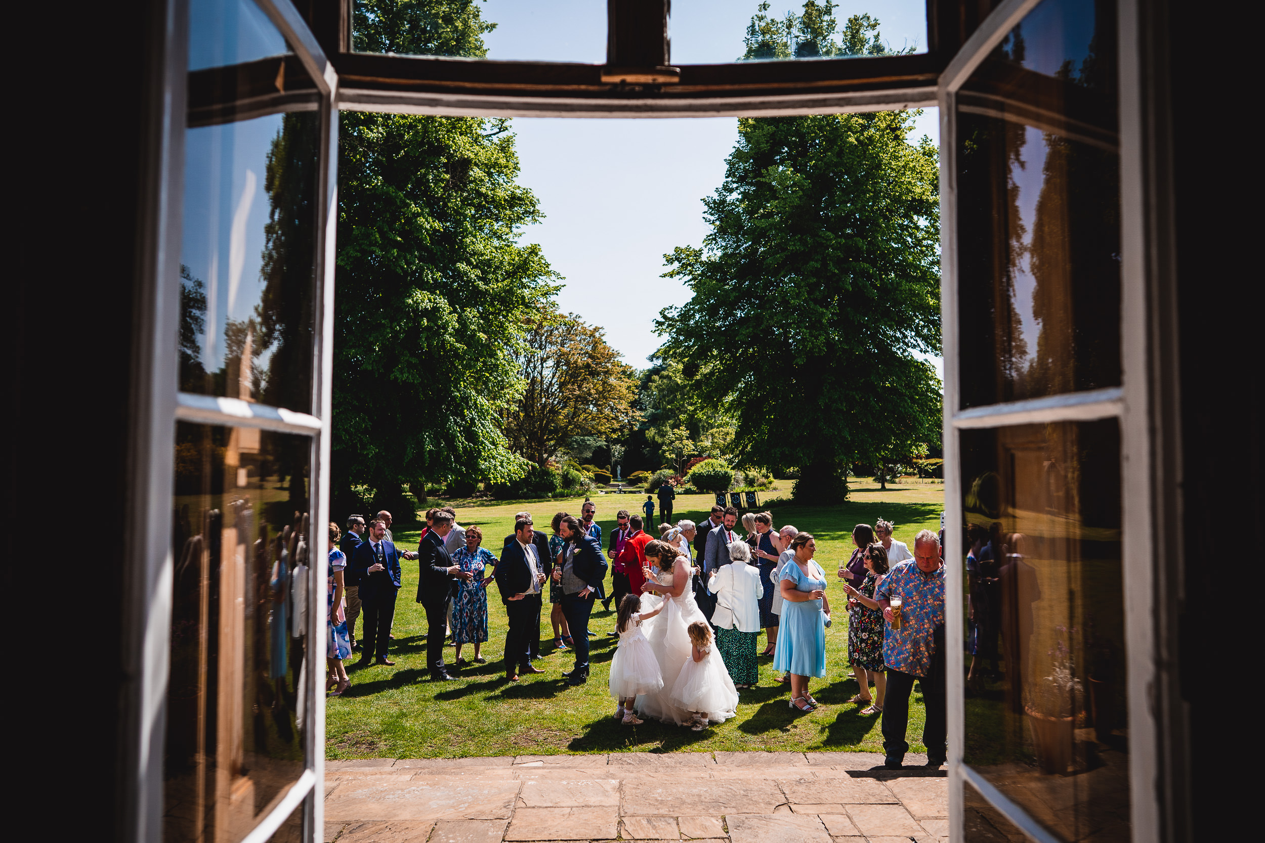 A group of people gathered outside in a garden setting on a sunny day, viewed through open double doors.