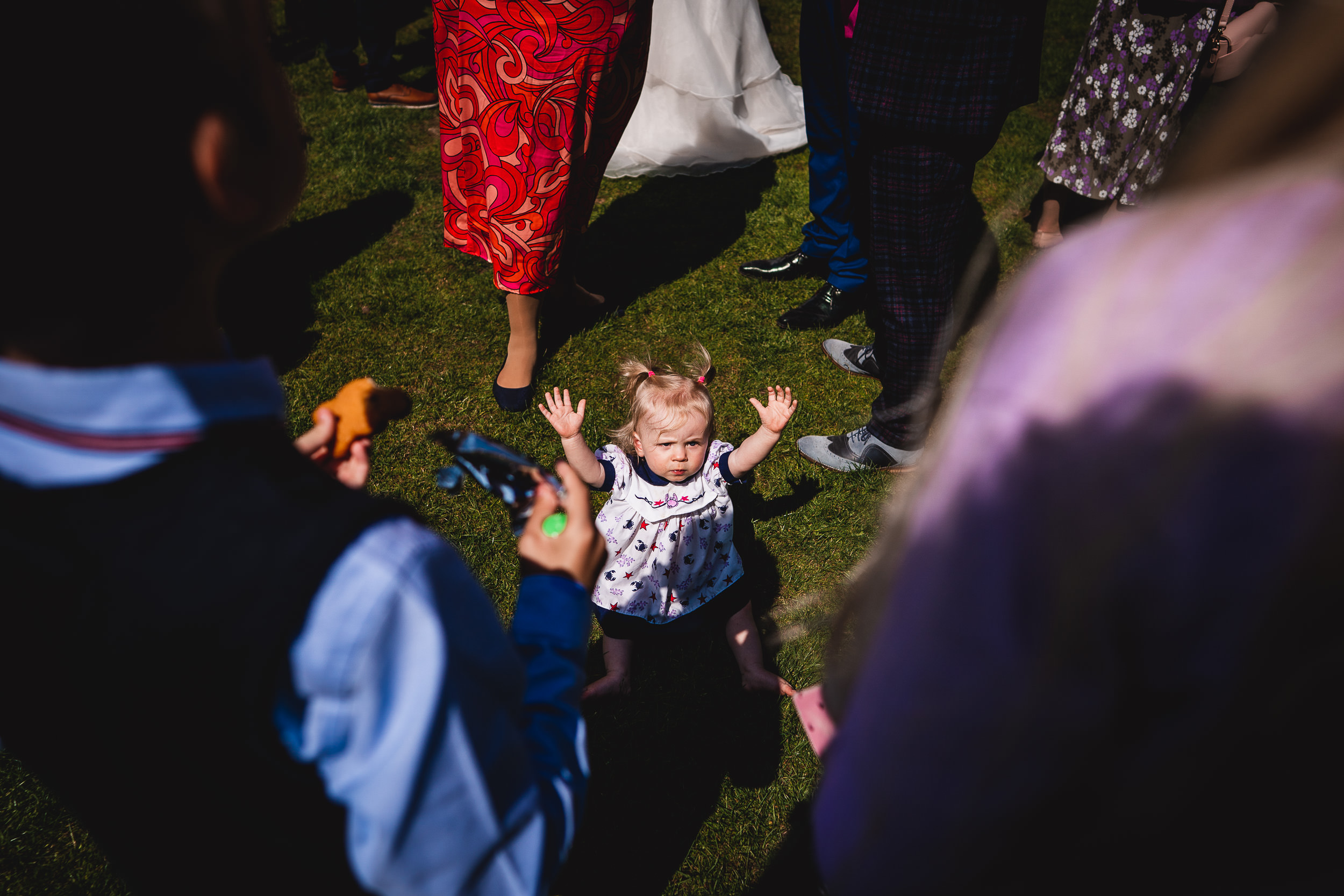 A toddler in a white dress with arms raised stands on grass surrounded by adults.