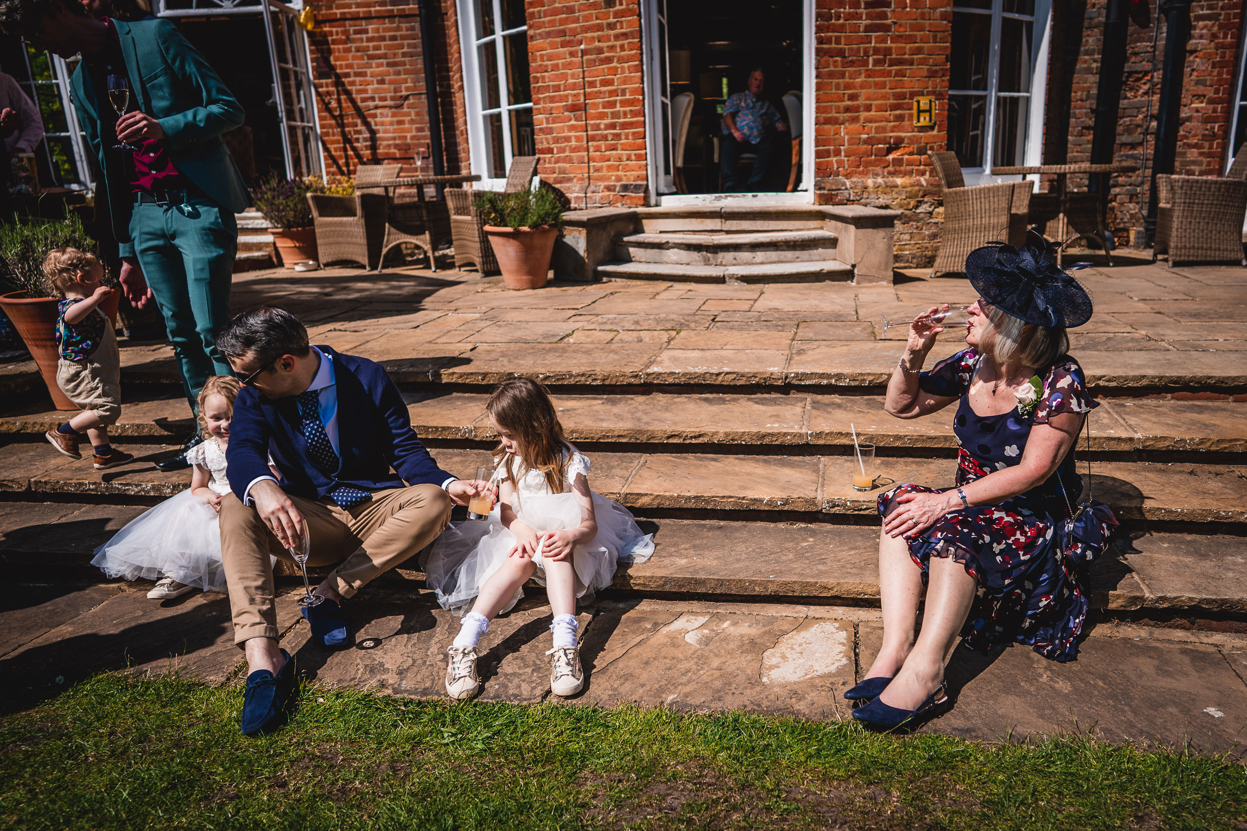 A group of people sit on steps enjoying a sunny day. An adult and child sit together, while another adult in a hat drinks from a glass. A child in the background walks with an adult.