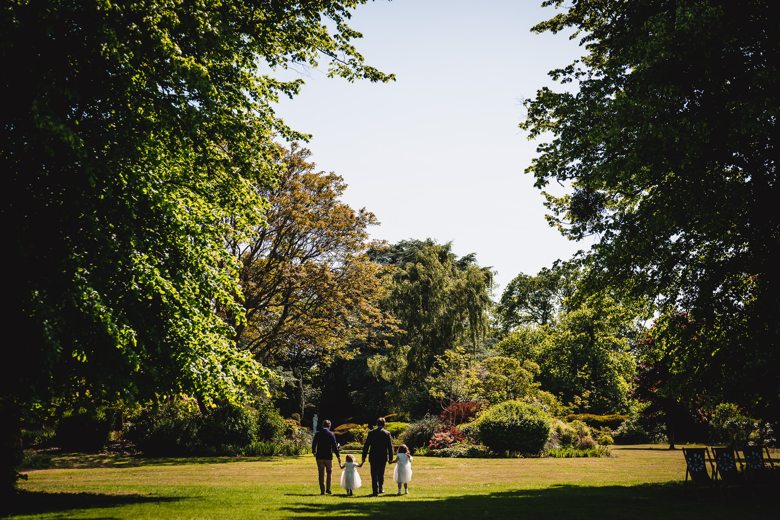 A man and three children walk hand-in-hand through a sunlit garden with trees and shrubs in the background.