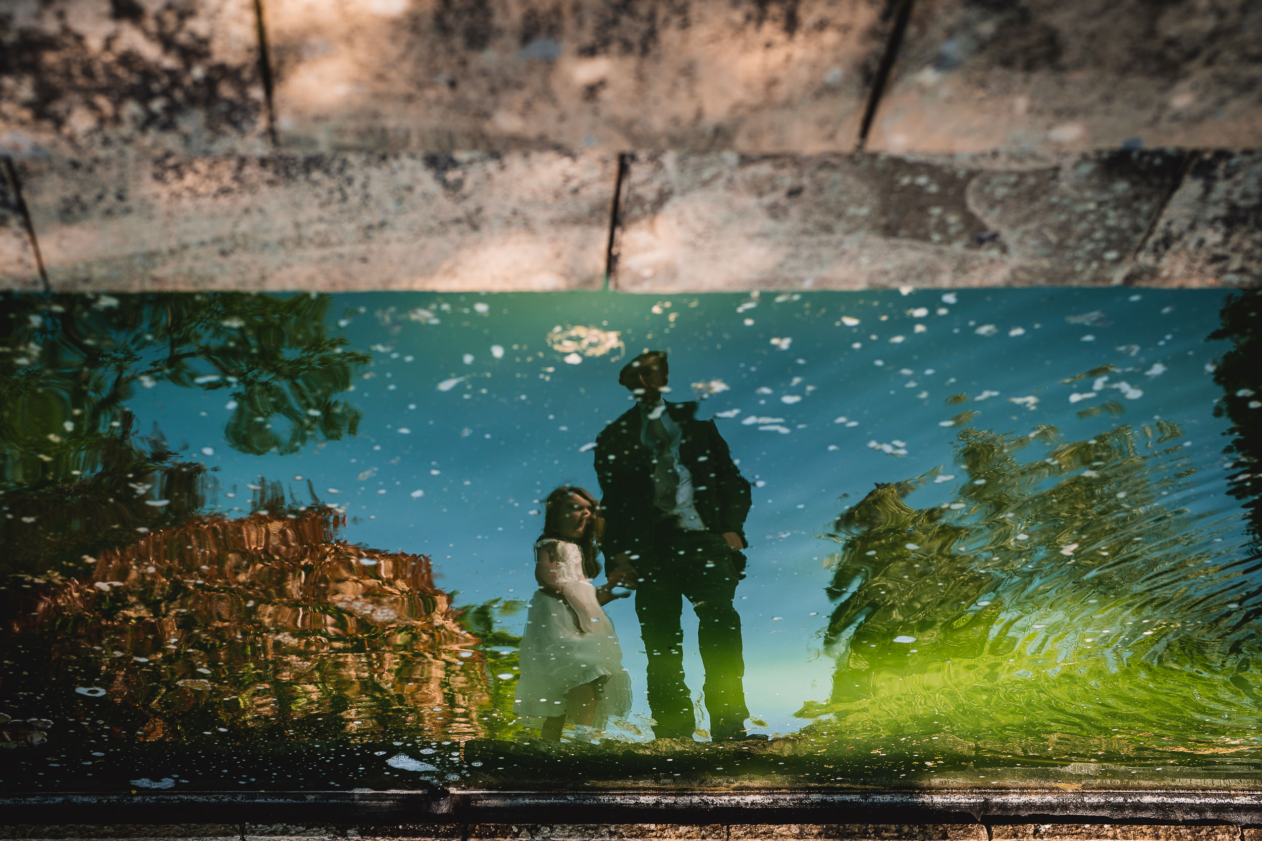 Reflection of a couple standing together, captured in a greenish water surface with surrounding trees and sky visible.