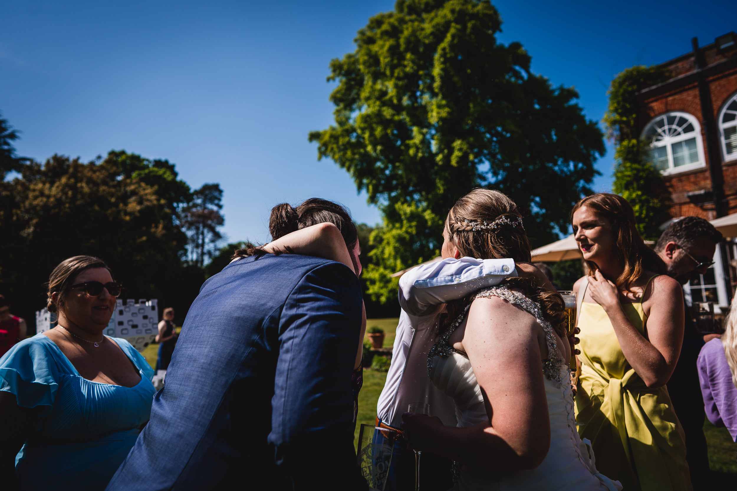 People in formal attire socializing outdoors at a gathering, with a large tree and a red brick building in the background under a clear blue sky.