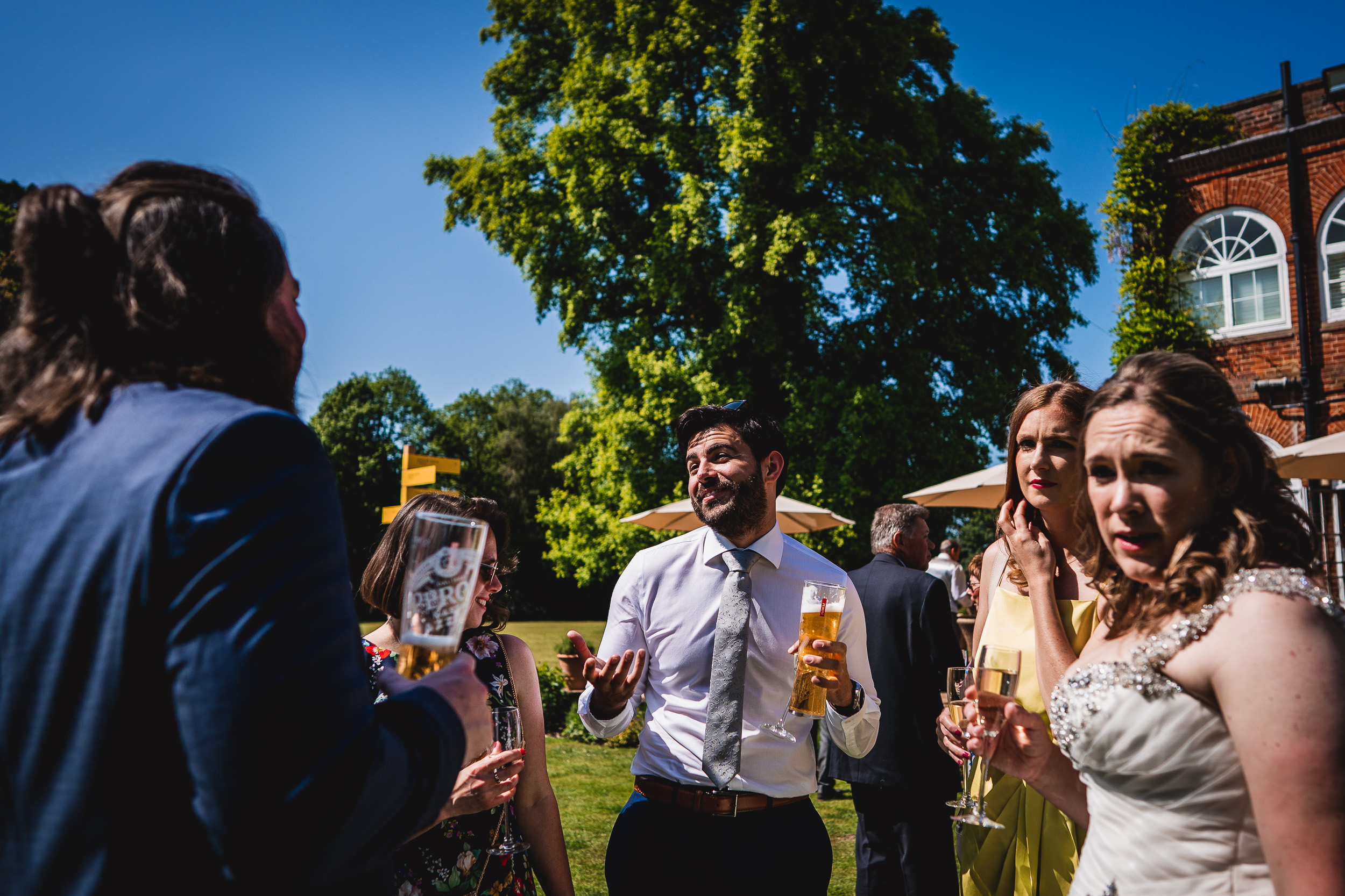 A group of people outdoors, dressed in formal attire, holding drinks and engaged in conversation on a sunny day with a large tree and a red brick building in the background.