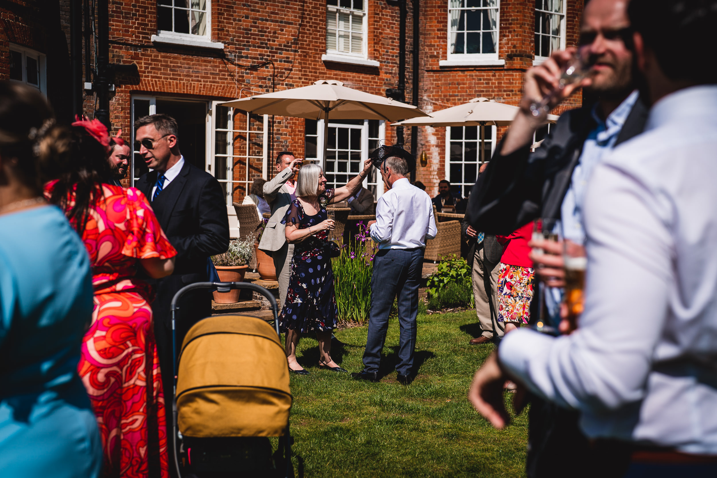 People socializing outdoors at a gathering, with some holding drinks. There is a stroller and umbrellas in the background, and a brick building is visible.