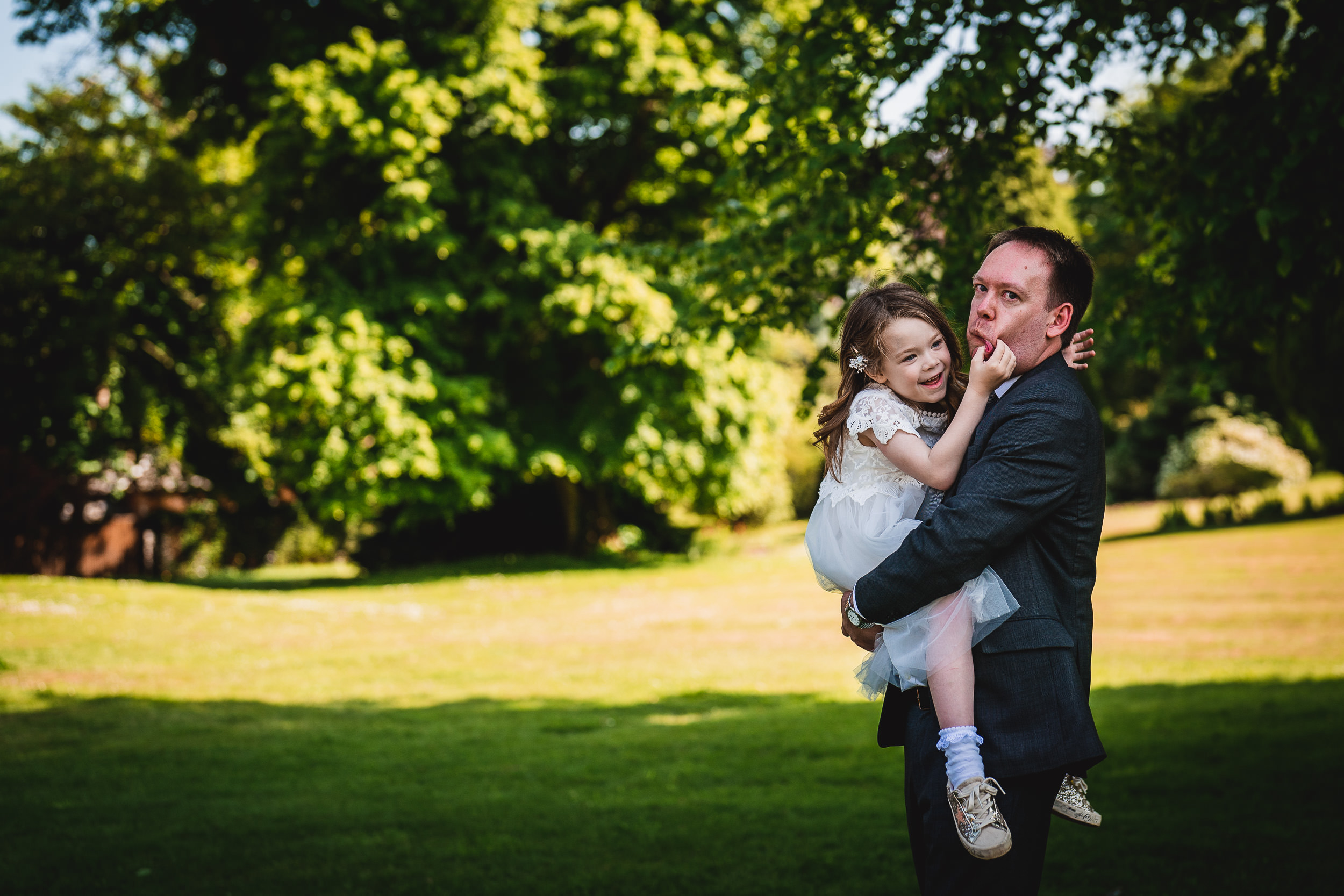 A man in a suit holds a smiling child in a white dress outdoors with green trees in the background.