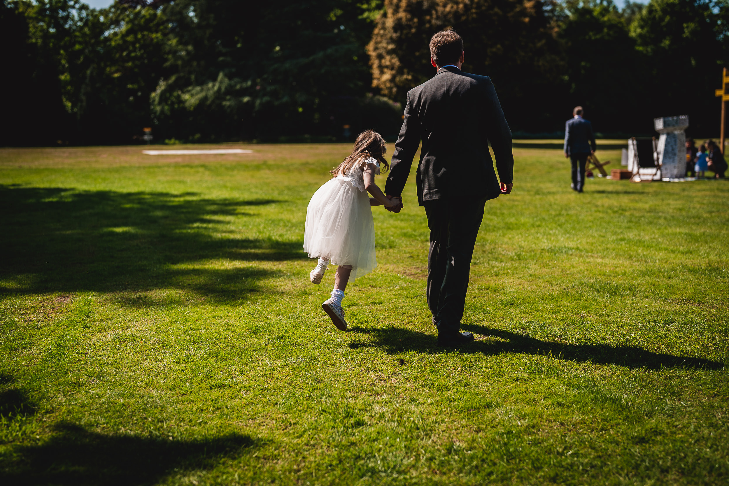 A man in a suit walks hand-in-hand with a young girl in a white dress across a sunny grassy field.