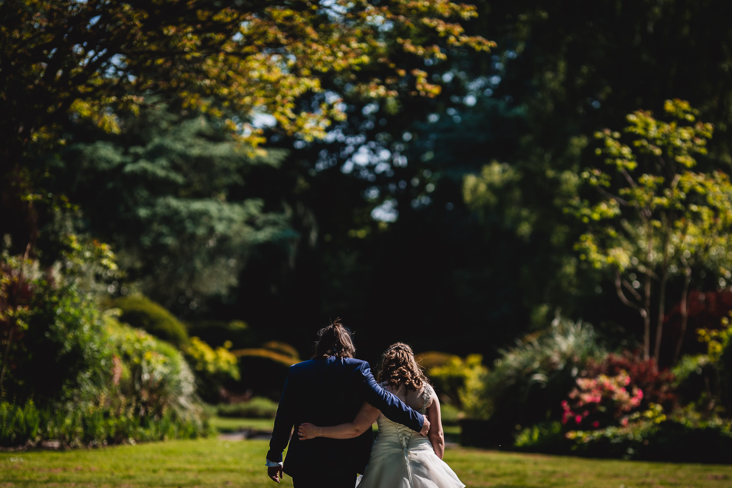 A couple in wedding attire walks arm in arm through a lush garden, surrounded by trees and colorful flowers.