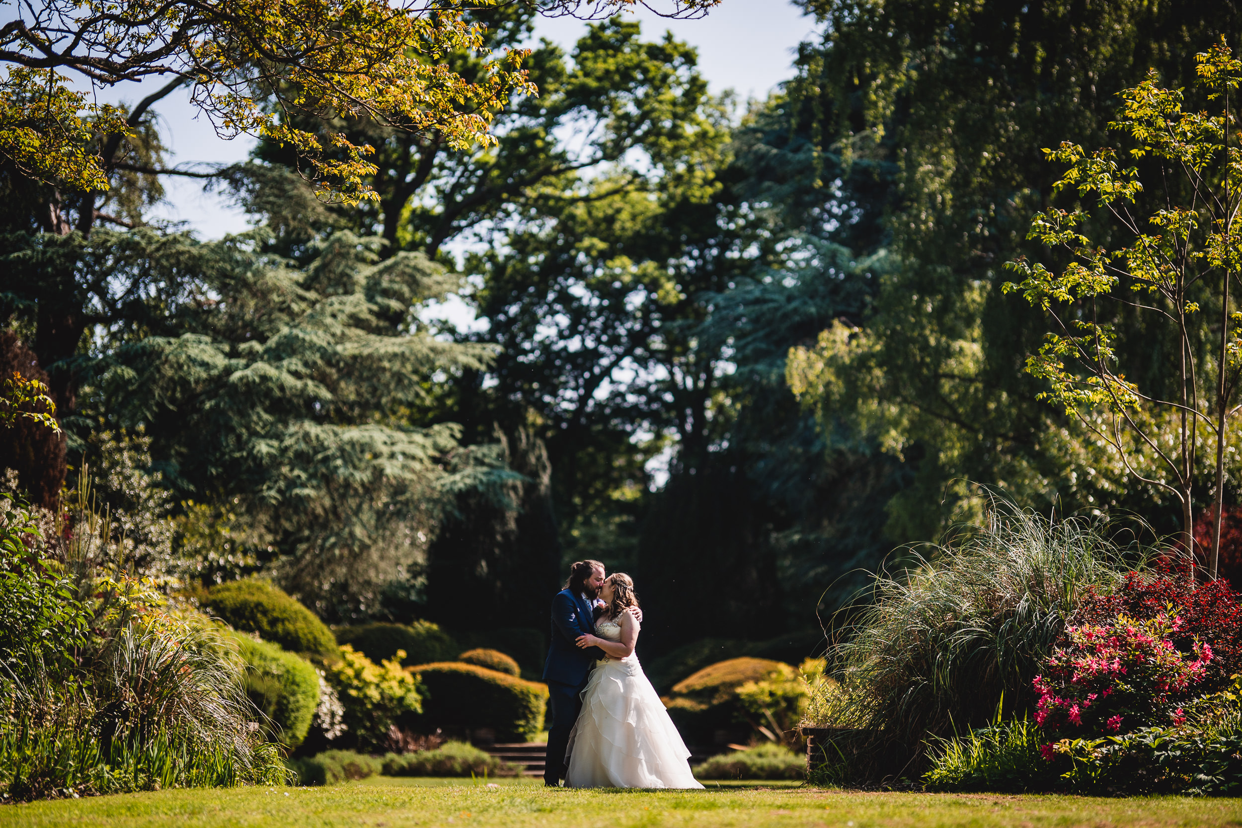 A bride and groom embrace in a lush, sunlit garden surrounded by trees and flowering plants.