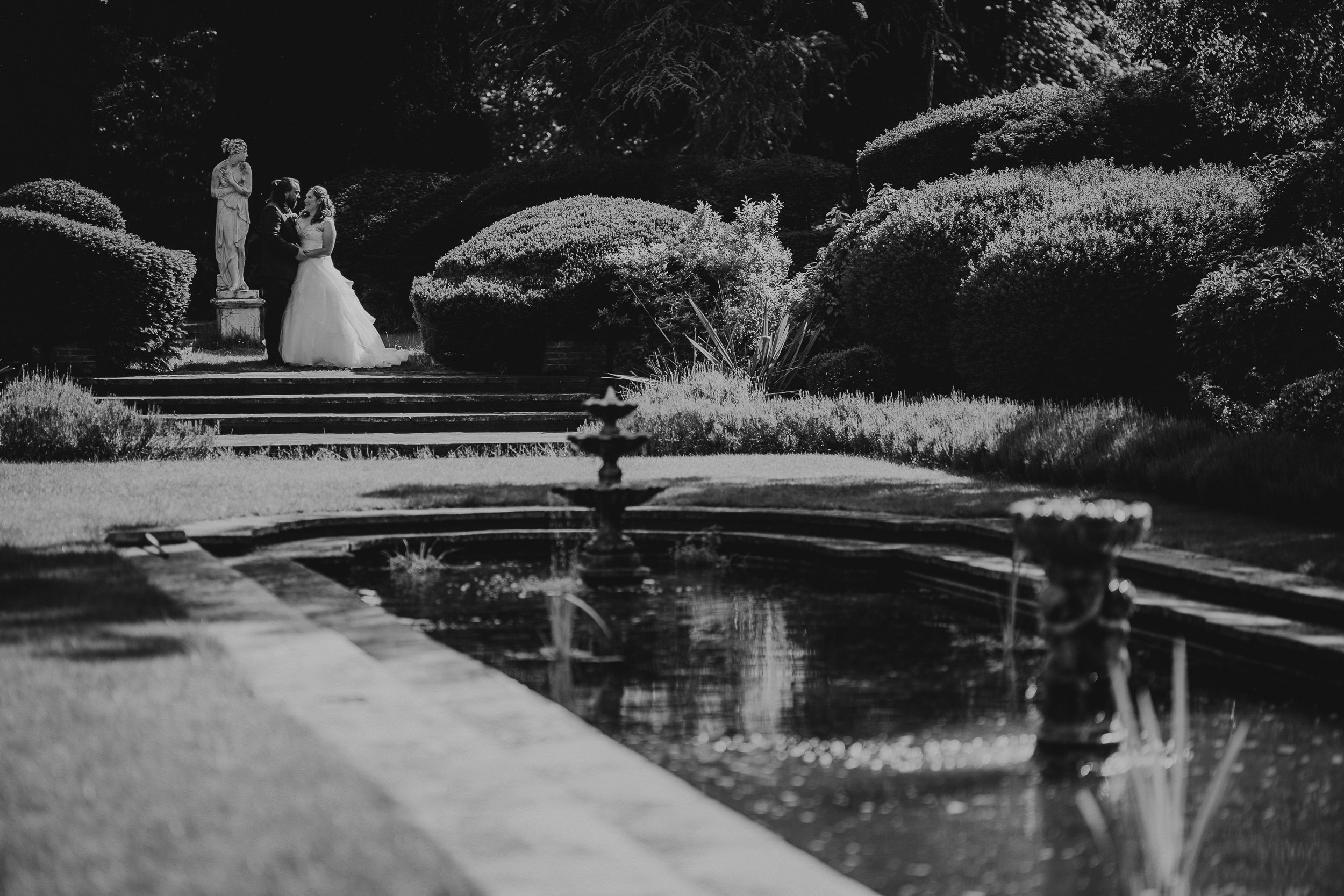 Bride and groom stand by a statue in a garden, with a reflecting pool in the foreground.