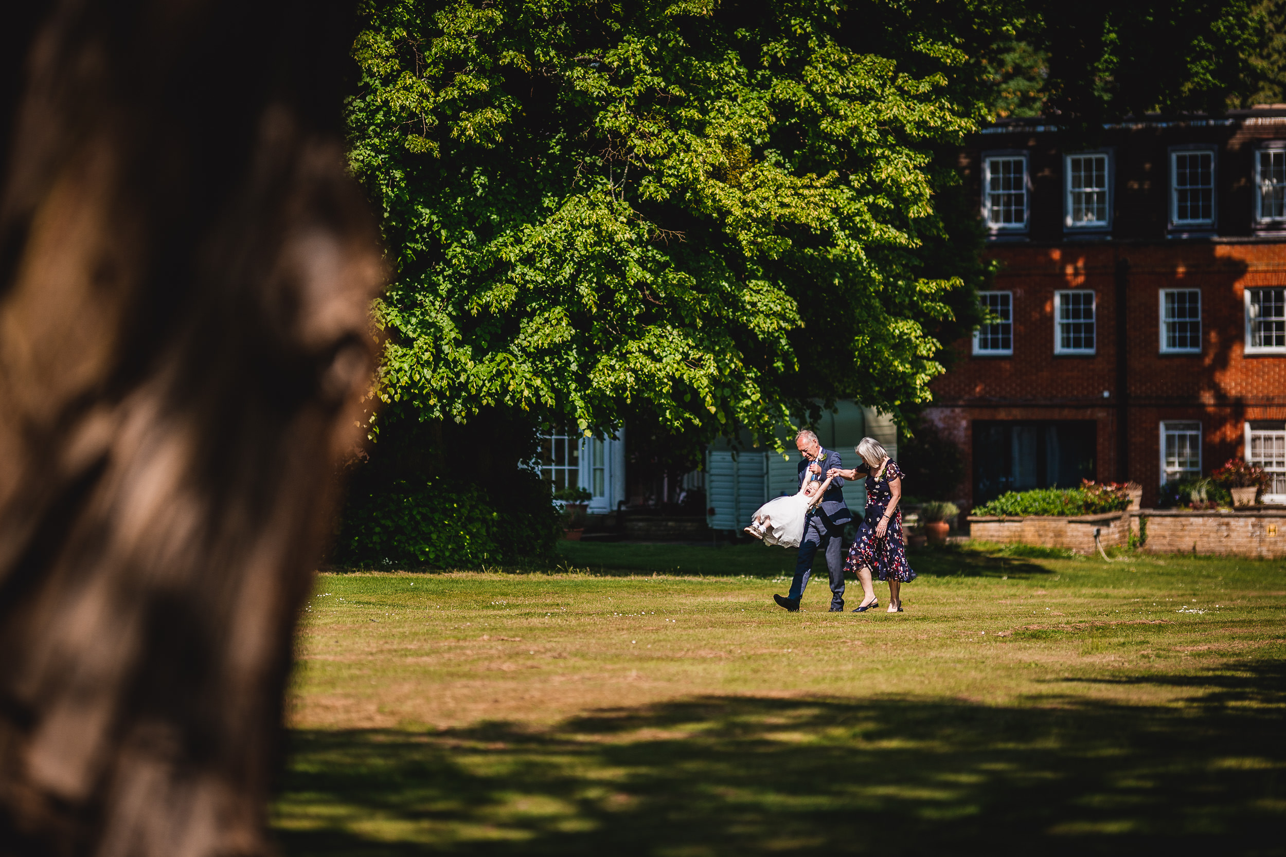 Two people walk together in a grassy area under a large tree with a brick building in the background.