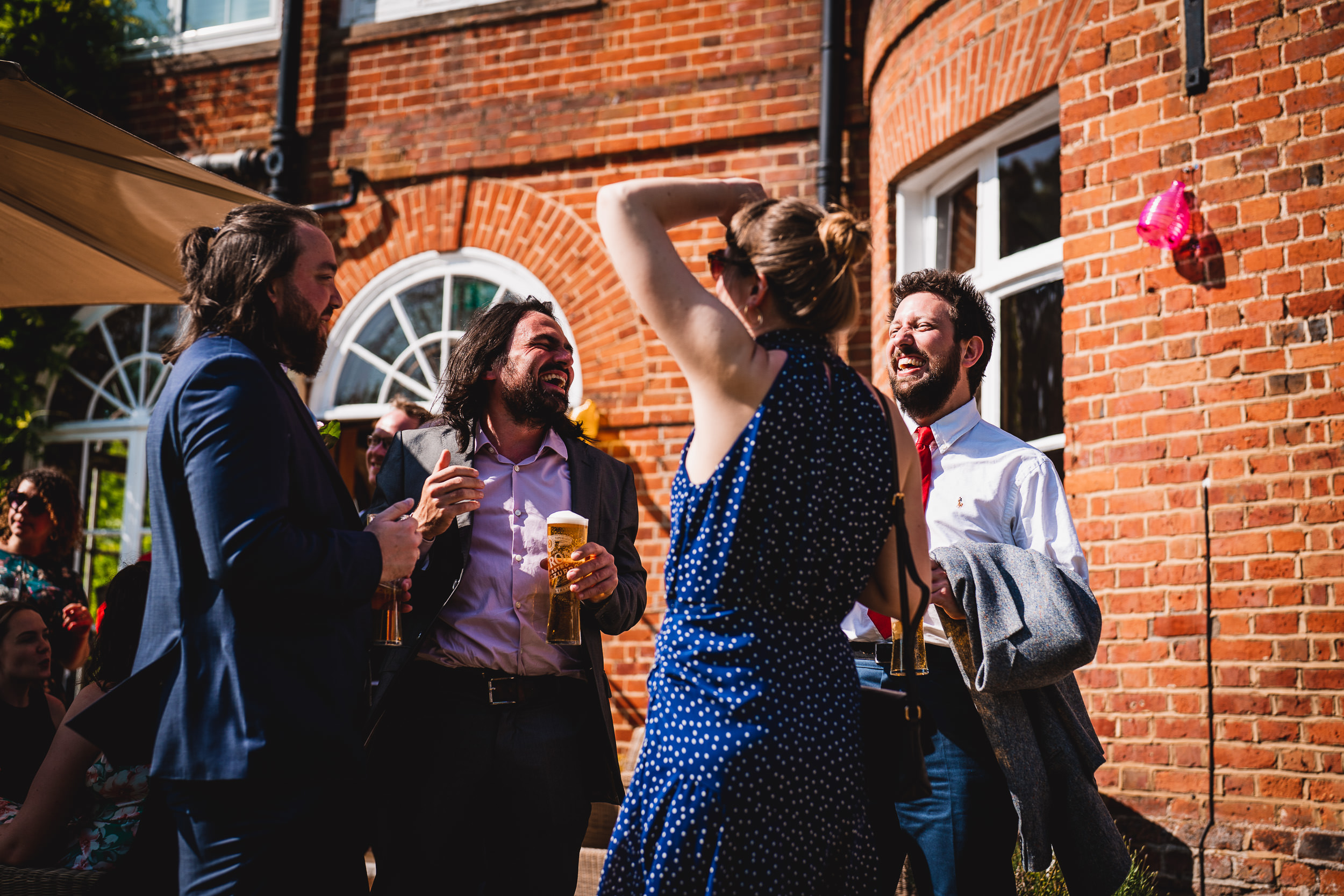 A group of people standing and laughing outdoors near a red brick building, with some holding drinks.