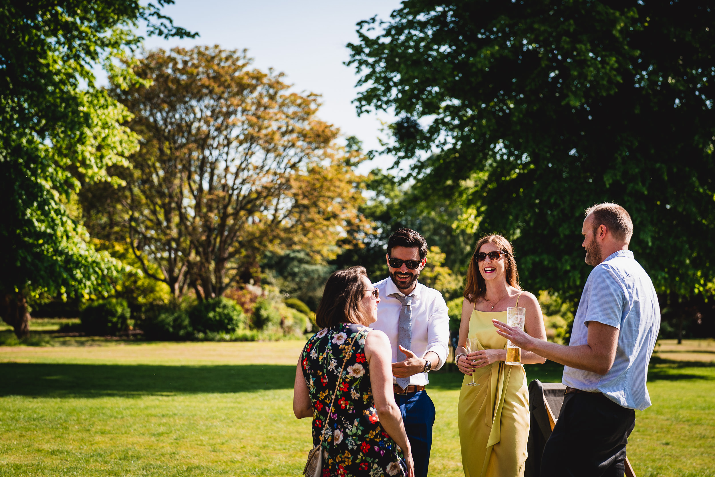 Four people converse and laugh outdoors in a sunny park, surrounded by trees and grass.