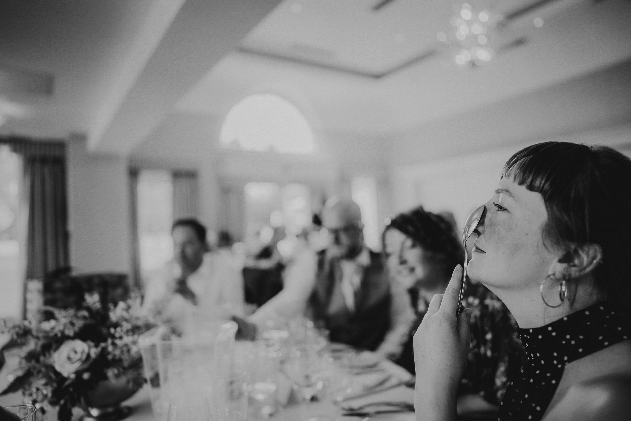 Black and white photo of people sitting at a dining table. A woman in the foreground looks thoughtful, with other guests blurred in the background. Elegant setting with soft lighting.