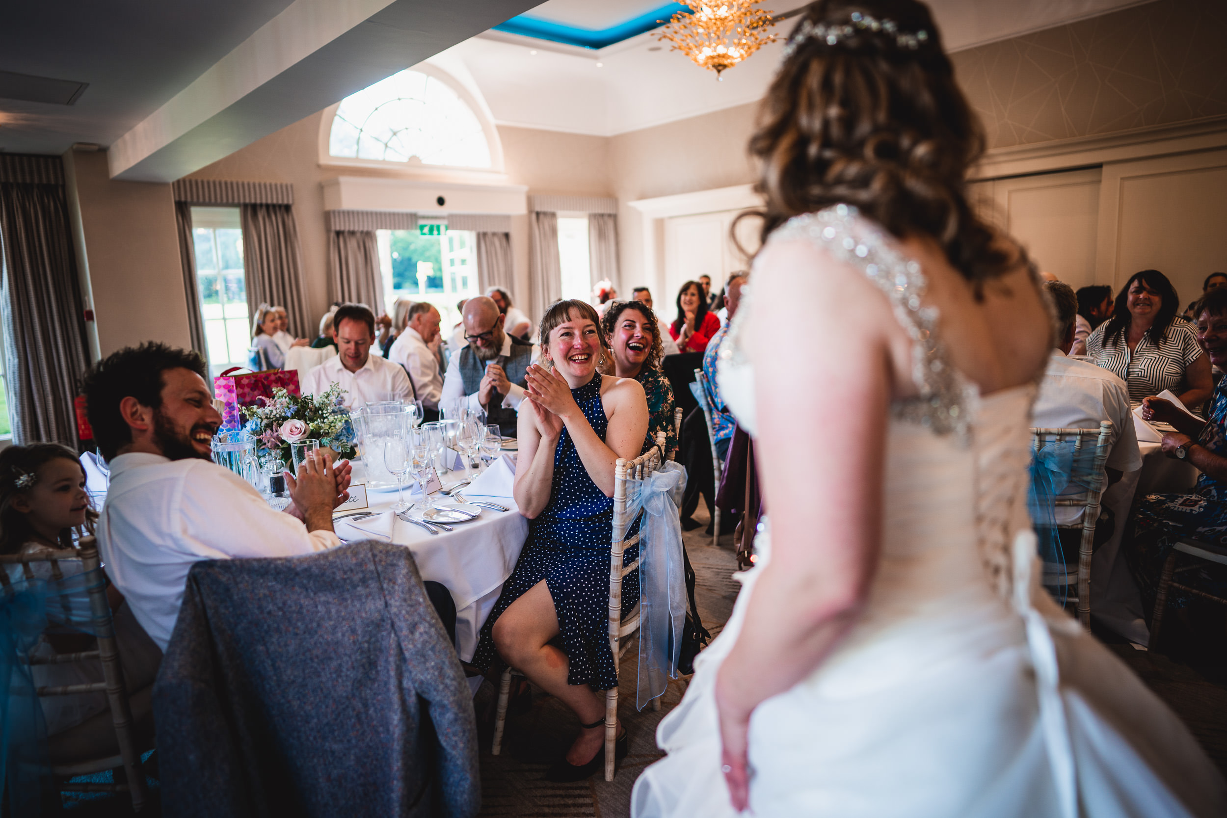 A bride stands in front of seated guests at a wedding reception. Guests are clapping and smiling in a bright, decorated room.