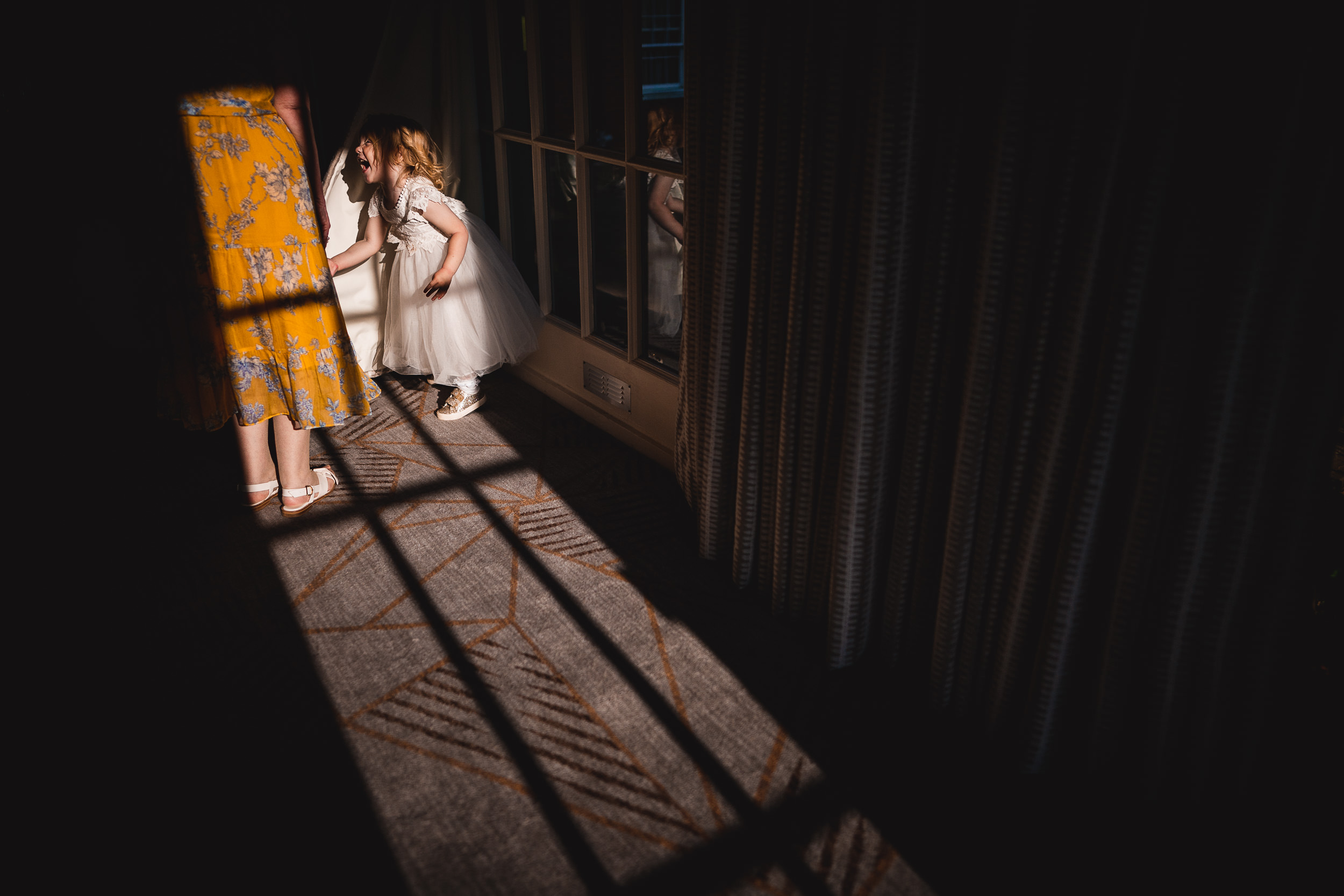 Two children in white dresses peek into a room through a partially open door. A person in a yellow floral dress stands nearby. Sunlight and shadows create patterns on the floor.