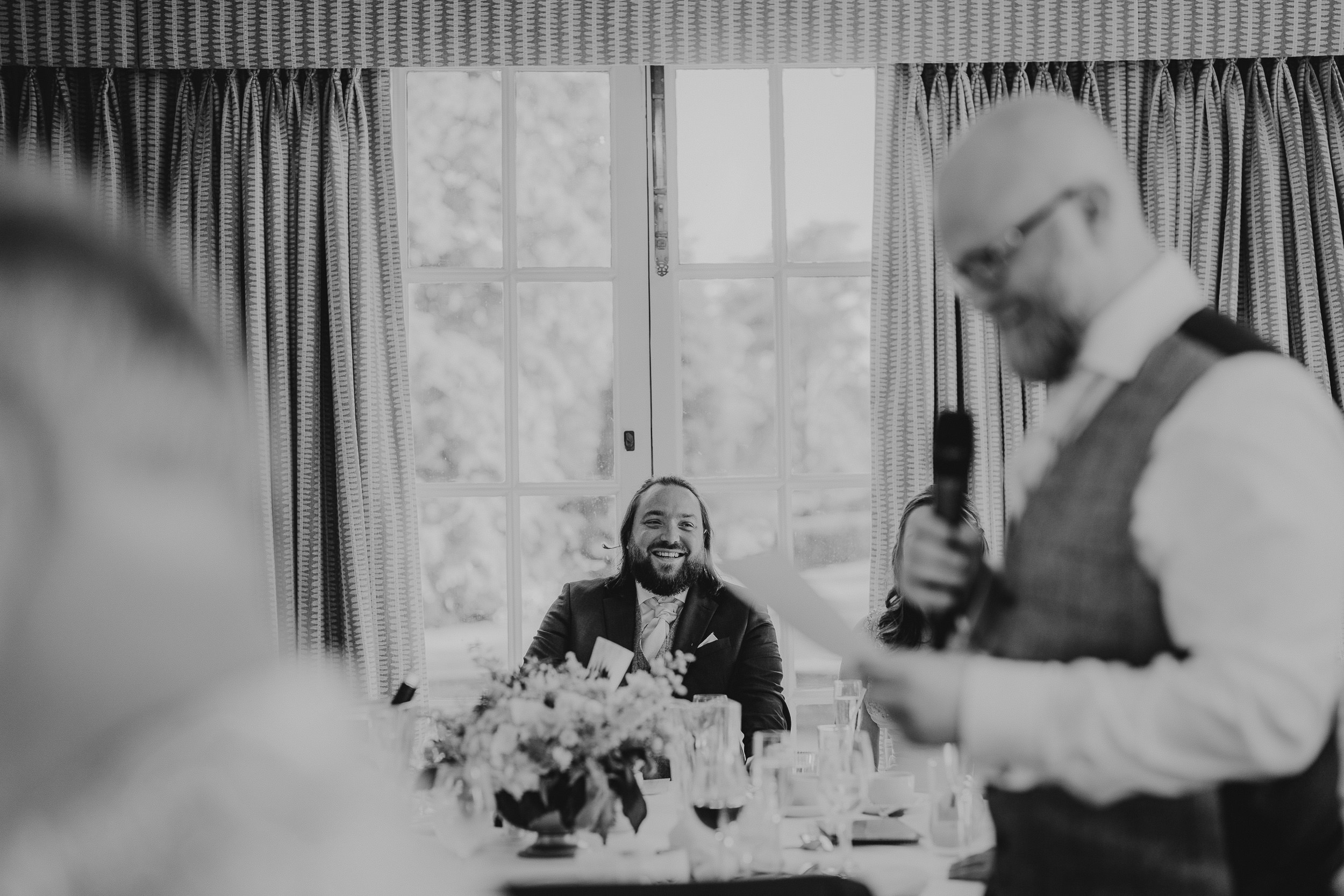 A man in a suit is seated at a table, smiling, while another man stands, holding a microphone and reading from a paper in a room with large windows and curtains.