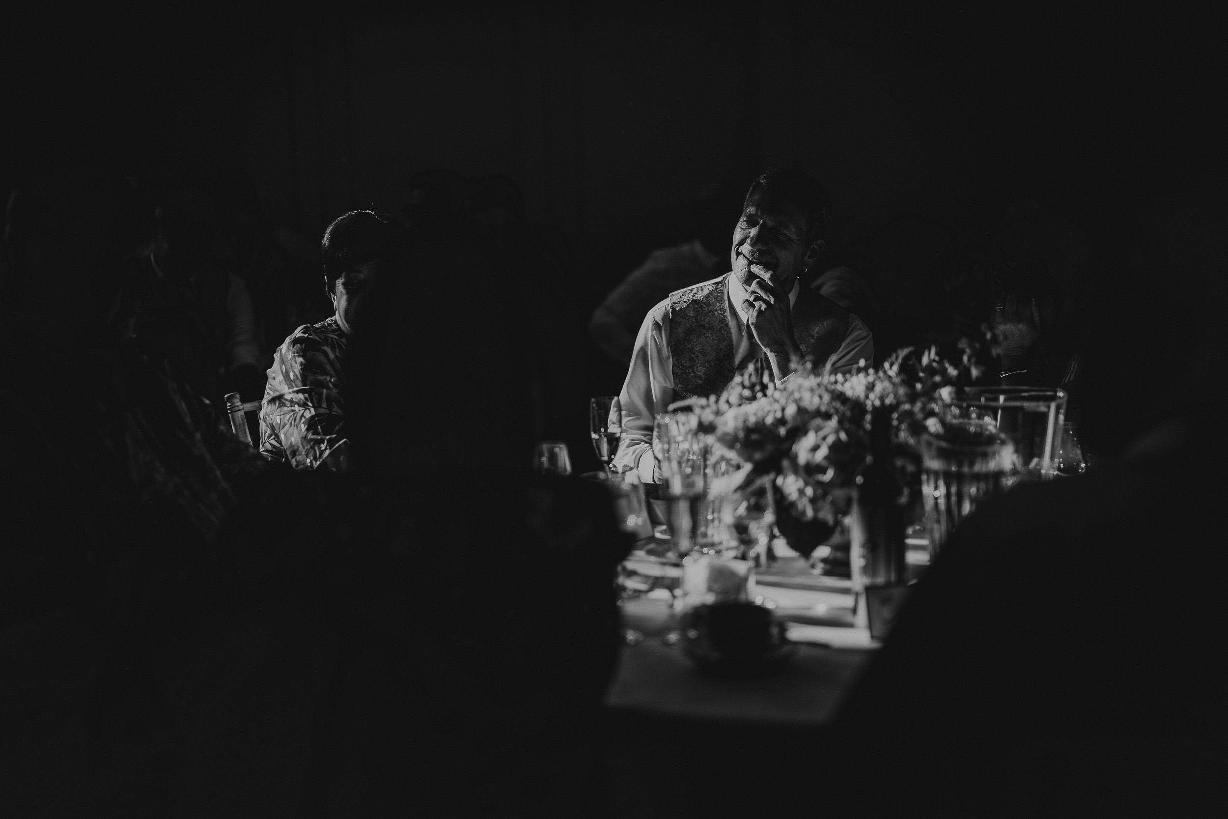 A person speaking into a microphone at a dimly lit event, sitting at a table with flowers and glassware.