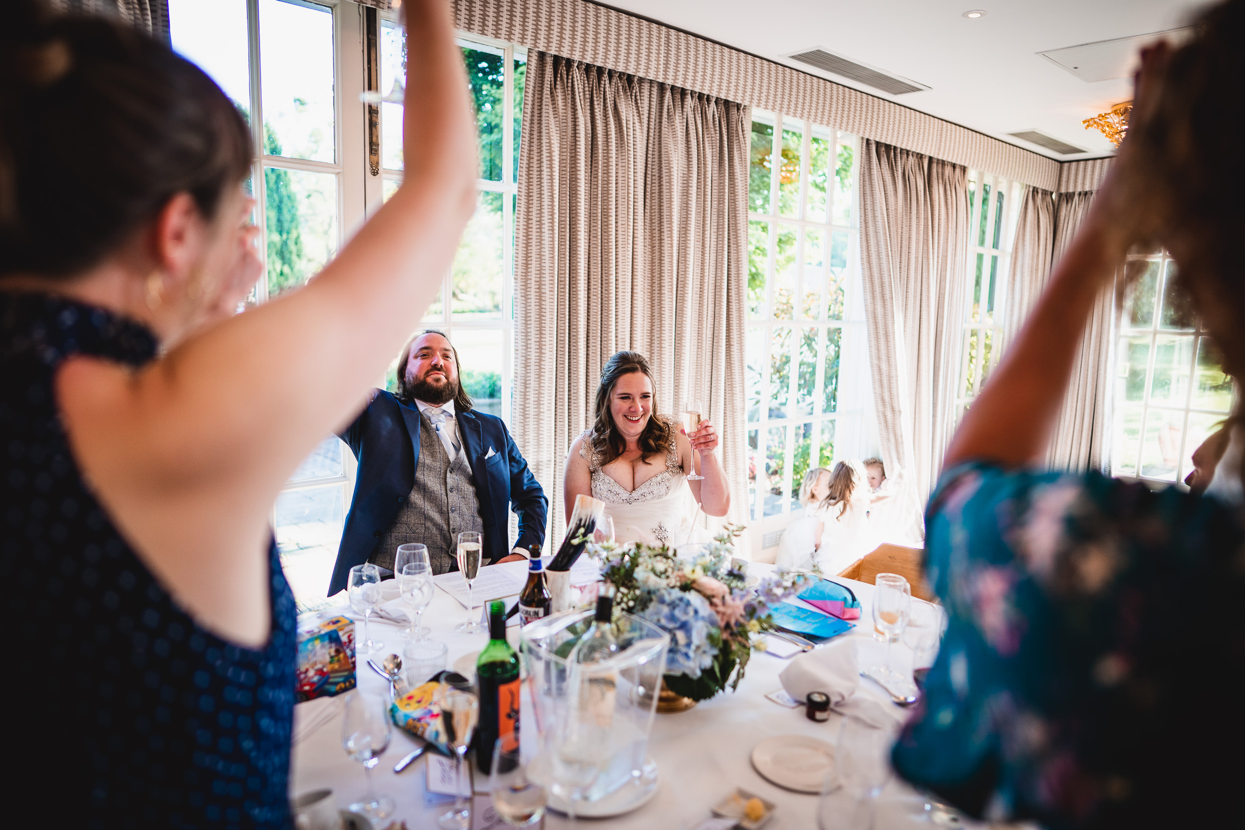 Bride and groom seated at a wedding reception table, surrounded by cheering guests. The room is well-lit with large windows in the background.