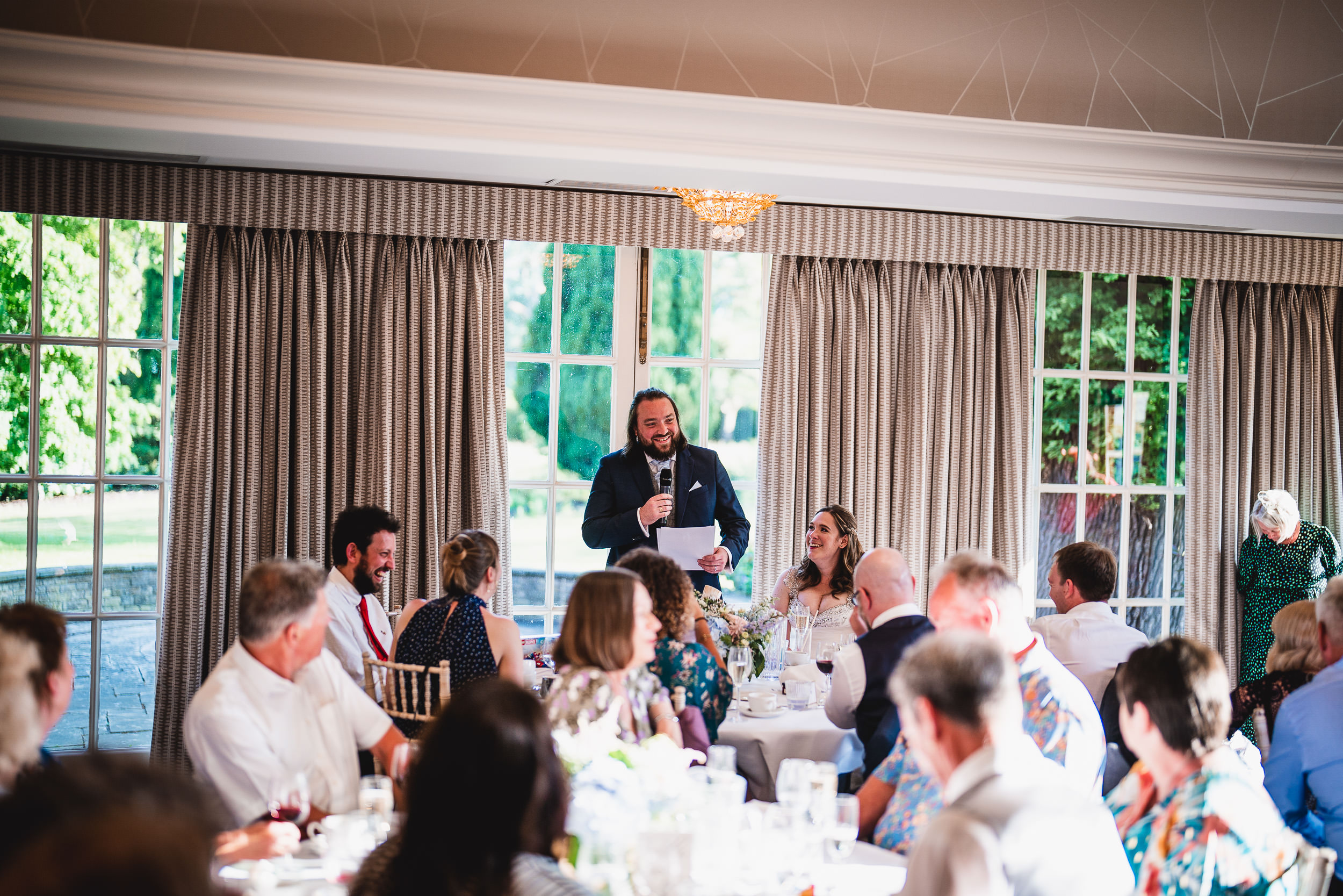 A man stands and speaks at a wedding reception, with seated guests and a smiling bride listening. Large windows in the background reveal a sunny, green outdoor scene.